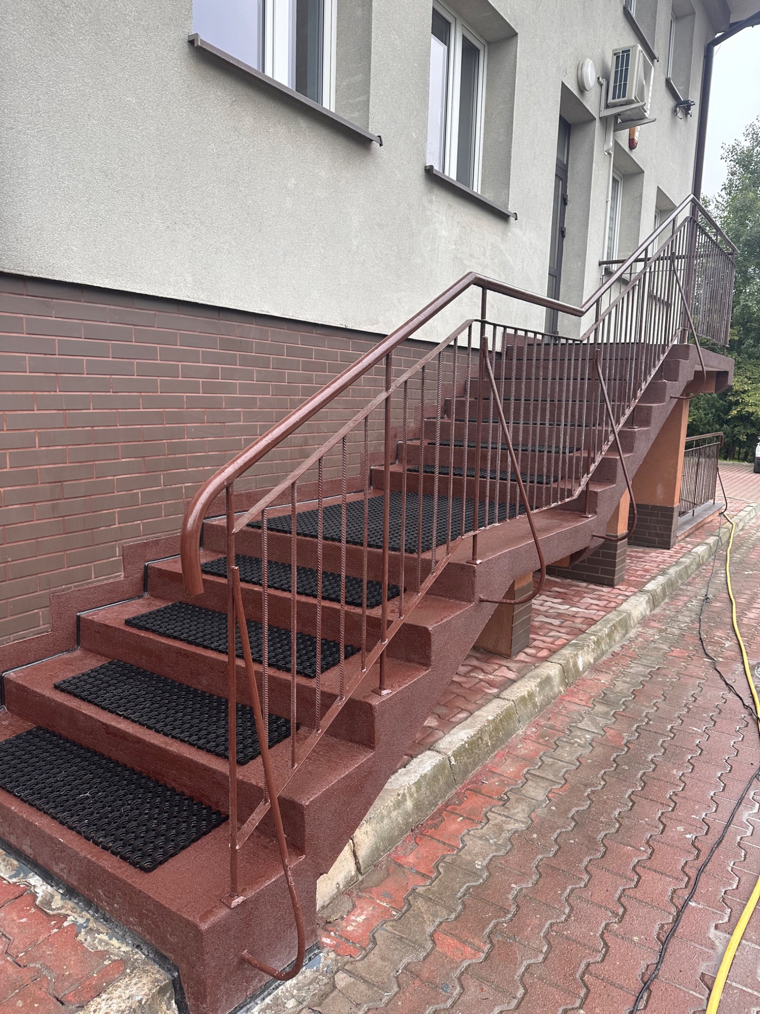 Zewnętrzne schody pokryte jednolitą, brązową warstwą, z czarnymi wycieraczkami na stopniach, prowadzące do budynku z szarą elewacją i brązową ceglaną podstawą.