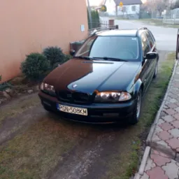 Czarny samochód kombi marki BMW zaparkowany przed budynkiem o pomarańczowej elewacji, widoczne krzewy i fragment chodnika z kostki.