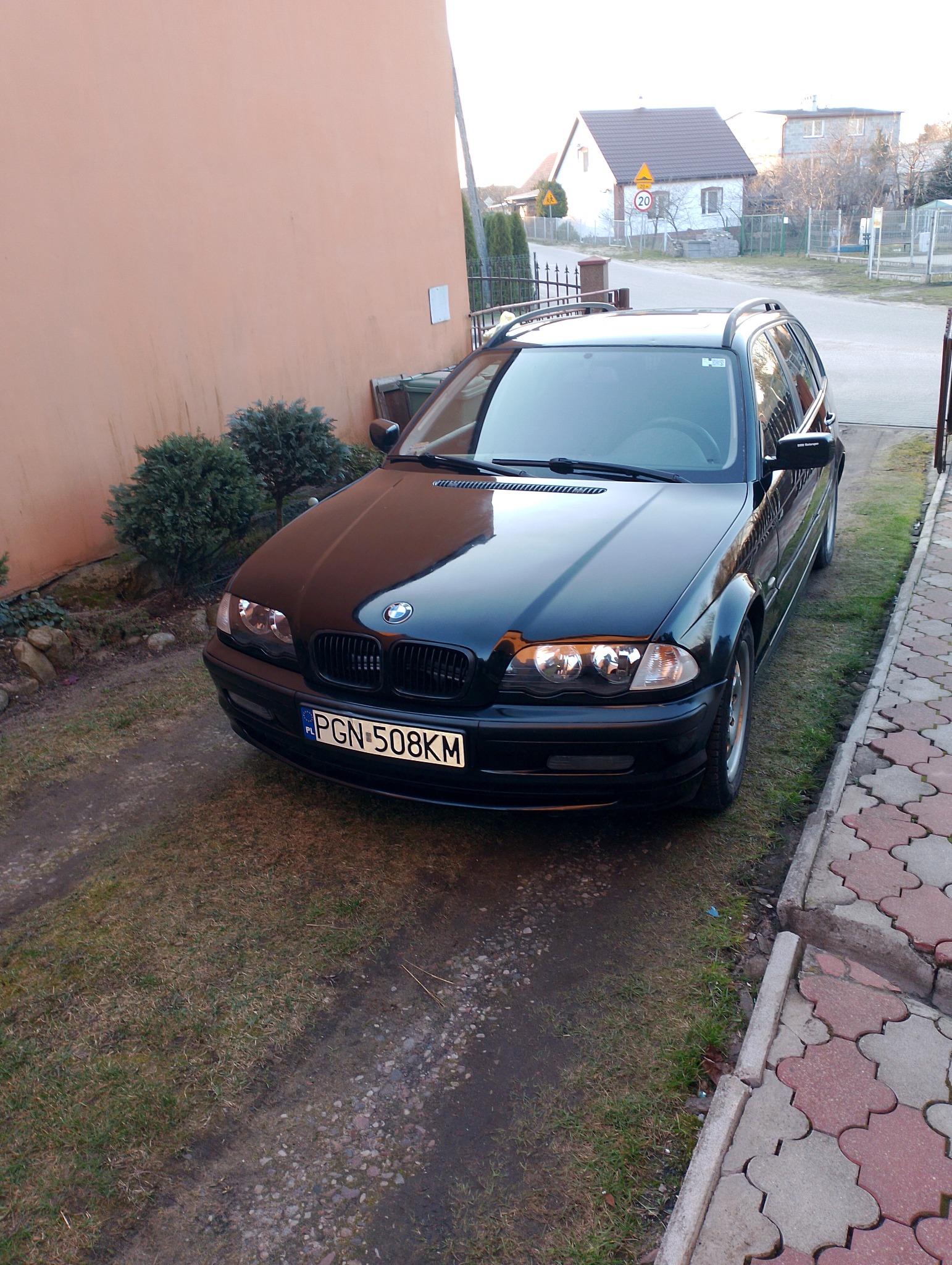 Czarny samochód kombi marki BMW zaparkowany przed budynkiem o pomarańczowej elewacji, widoczne krzewy i fragment chodnika z kostki.