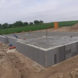 Wylany fundament z szalunkami traconymi na tle zielonego pola i pochmurnego nieba.