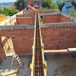 Wznoszenie ścian z czerwonej cegły ceramicznej na fundamencie, widok z g&oacute;ry na zbrojenie w szalunku, drabina i piła łańcuchowa na posadzce, w tle zabudowa jednorodzinna.