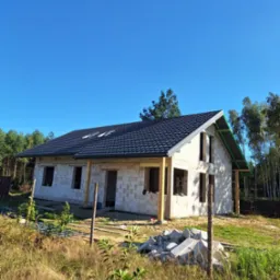 Stan surowy otwarty domu z białych bloczków, czarny dach z oknami dachowymi, drewniane podparcie dachu, materiały budowlane na trawie, błękitne niebo.