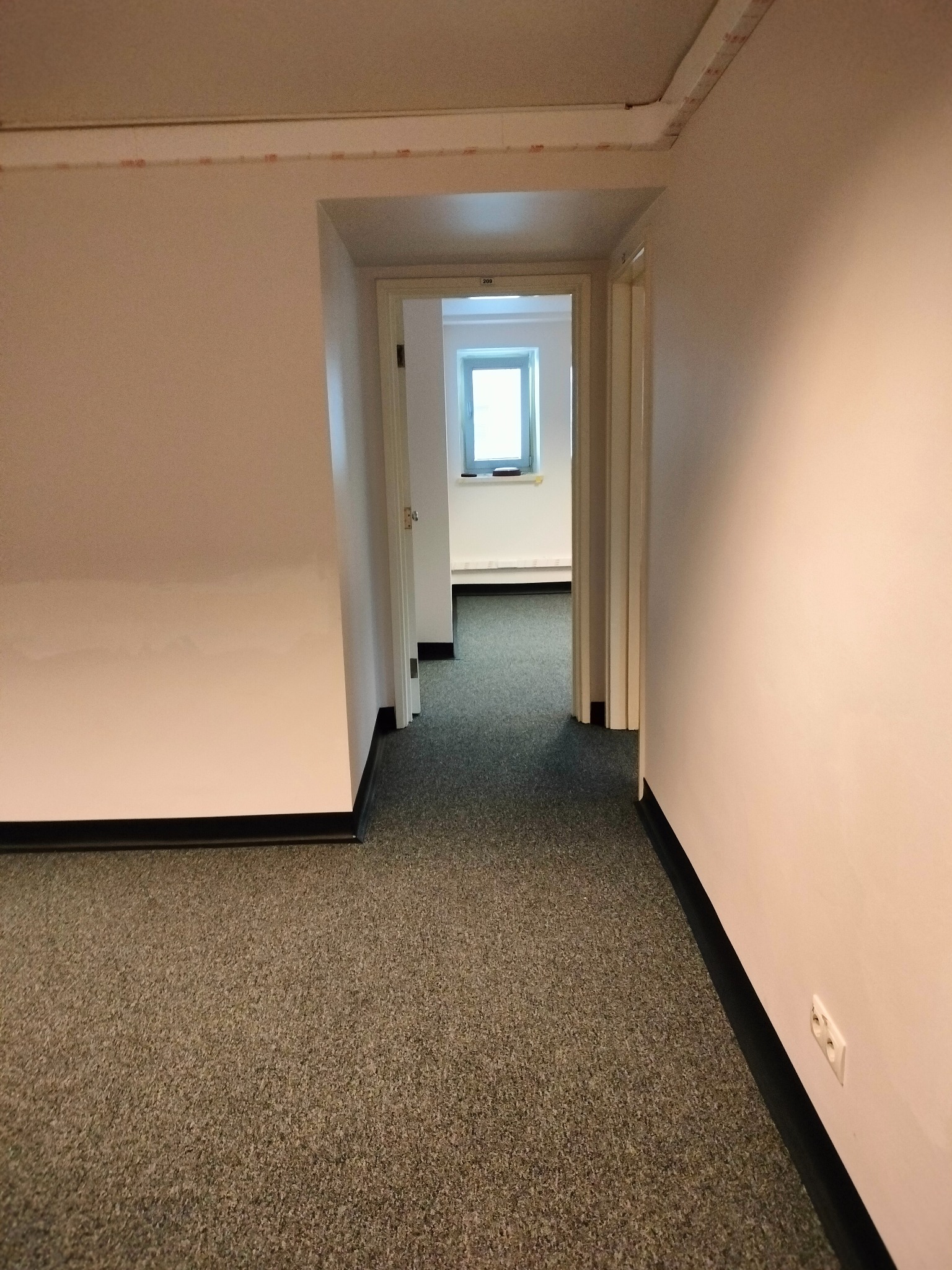 Korytarz biurowy z szarą wykładziną dywanową, pomalowanymi na jasny kolor ścianami i widocznymi gniazdkami elektrycznymi oraz drzwiami w głębi prowadzącymi do kolejnych pomieszczeń.