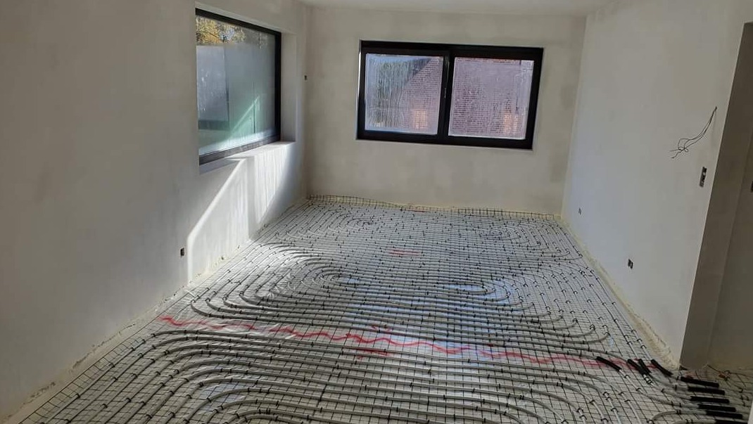 Białe, surowe wnętrze z widoczną instalacją ogrzewania podłogowego na siatce, przygotowane do wylewki, z dwoma oknami w czarnych ramach.