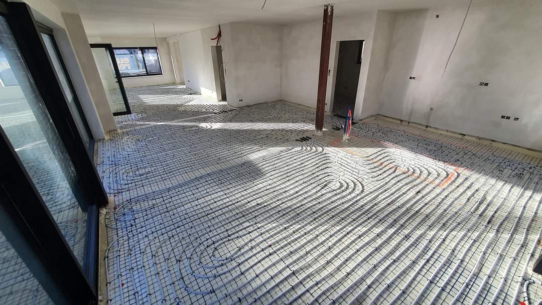 Białe rury ogrzewania podłogowego zamontowane na siatce w surowym pomieszczeniu z otworami okiennymi i drzwiami, widoczne wystające przewody i instalacje elektryczne.