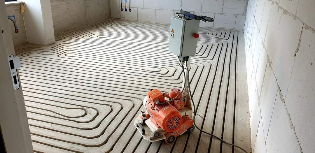Rozłożone rury ogrzewania podłogowego w surowym wnętrzu, widoczna pomarańczowa maszyna do wylewki i skrzynka sterownicza na podłodze z betonu komórkowego.