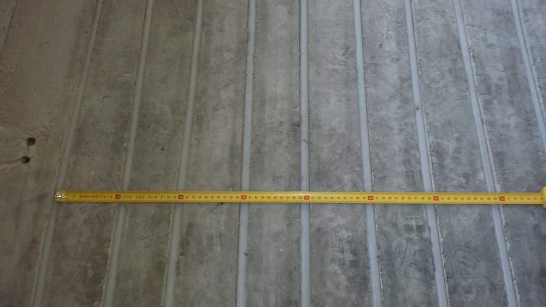 Szary betonowy podkład pod ogrzewanie podłogowe z widocznymi rowkami wypełnionymi szarym uszczelniaczem; żółta miarka rozłożona w poprzek.