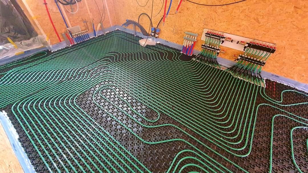 Zielone rurki ogrzewania podłogowego ułożone na czarnej macie z widocznymi rozdzielaczami na ścianie z płyty OSB.