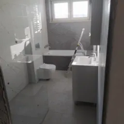 Wnętrze łazienki w trakcie wykańczania, widoczna kabina prysznicowa ze szklaną ścianą, umywalka z szafką, toaleta, wanna oraz drabina malarska.