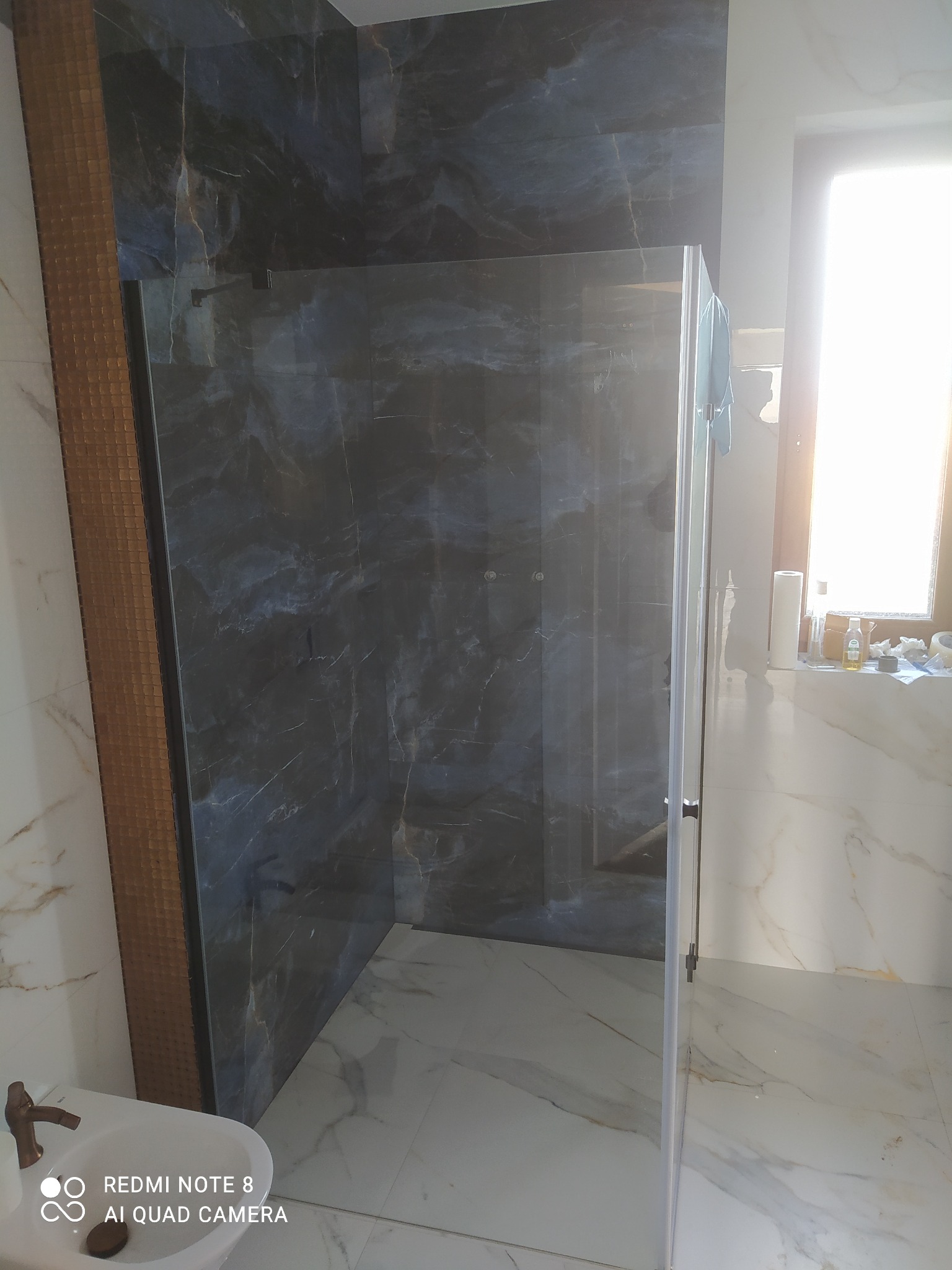 Nowoczesna kabina prysznicowa z czarnymi płytkami imitującymi marmur, złote akcenty na płytkach i armaturze, jasne marmurowe płytki podłogowe, widoczna toaleta w rogu.