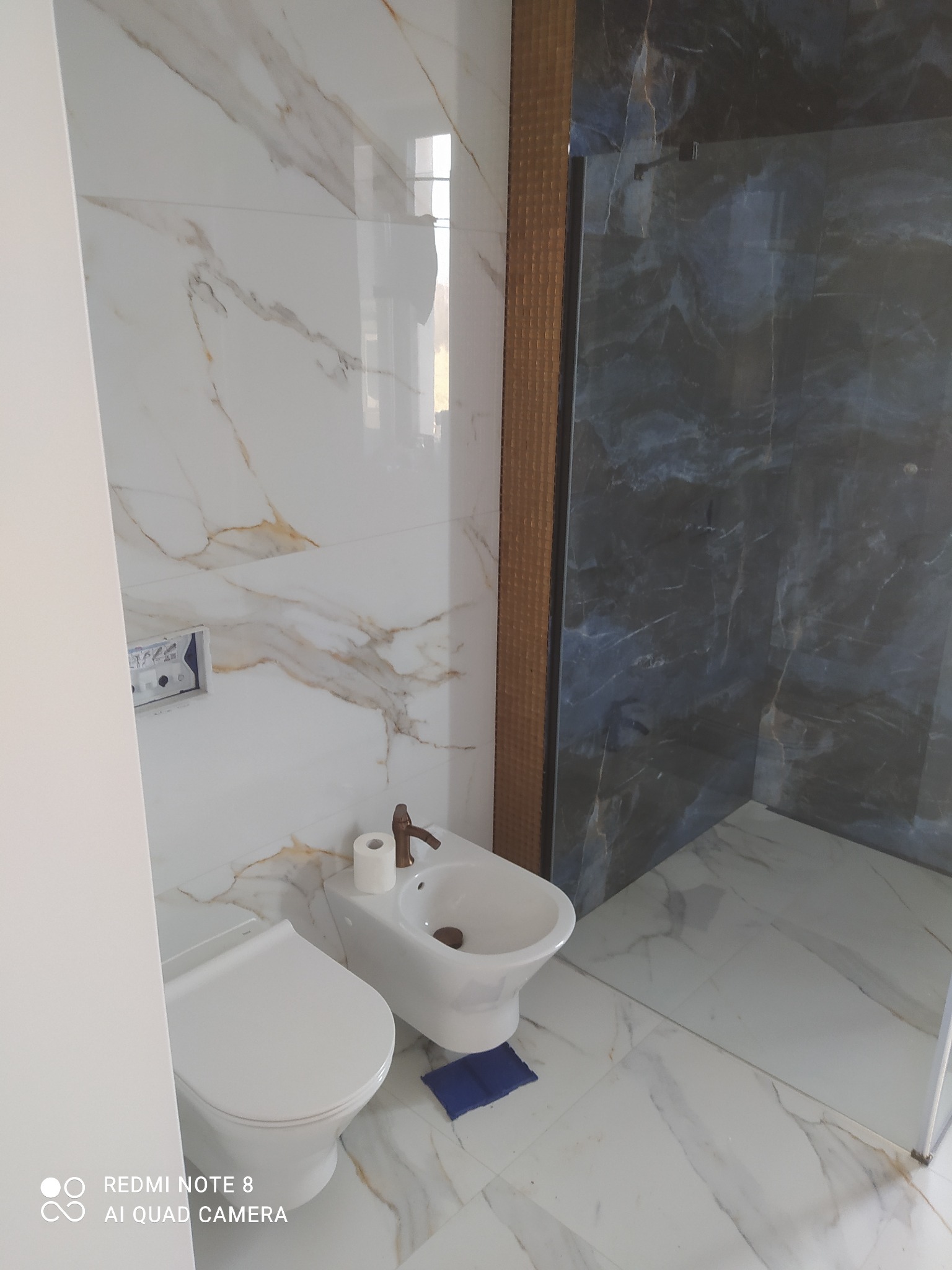 Wykończona łazienka z białymi marmurowymi płytkami, toaletą i bidetem obok kabiny prysznicowej z ciemnymi płytkami imitującymi marmur, widoczny detal w postaci małego niebieskiego dywanika...