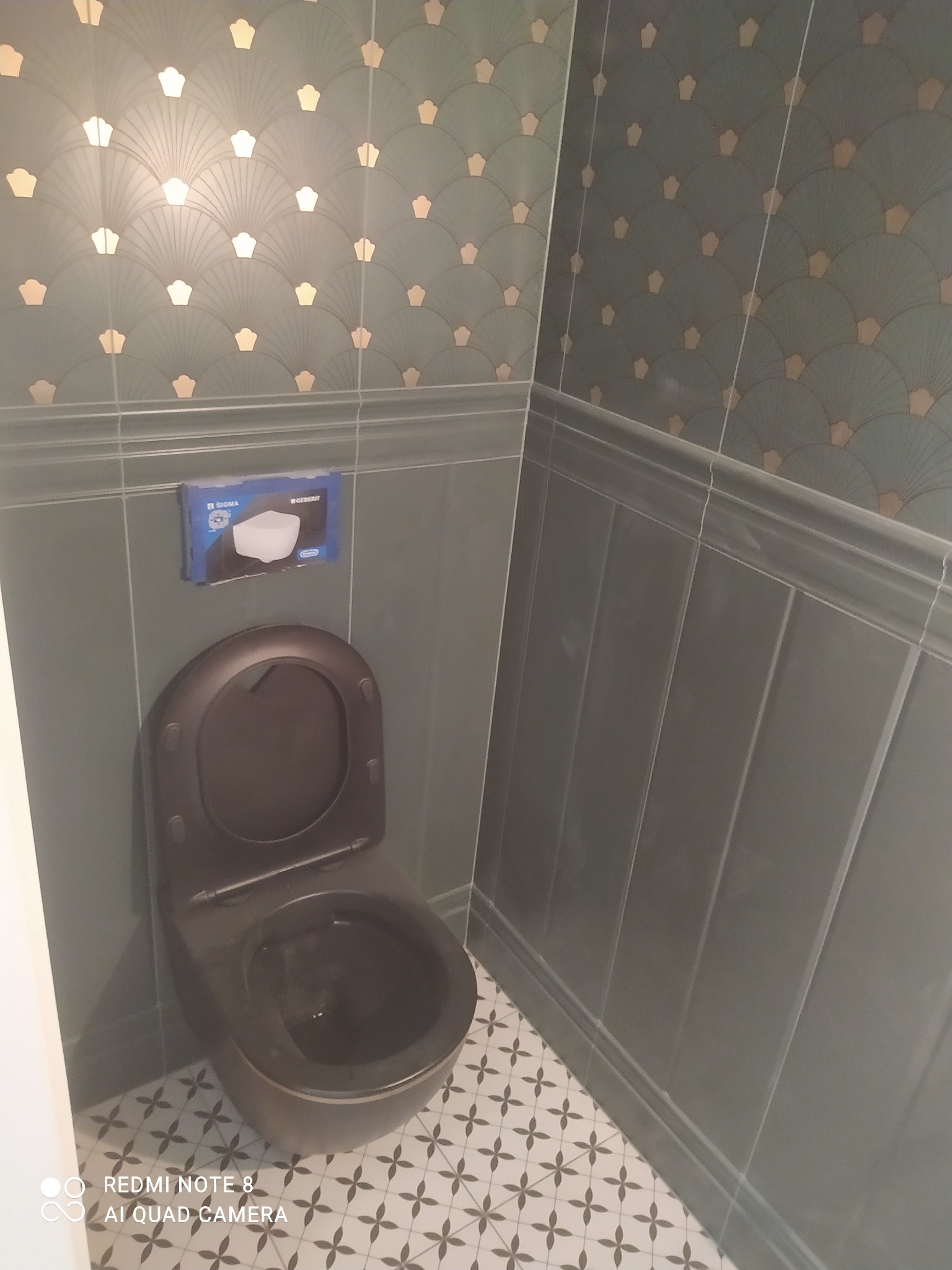 Zamontowana czarna toaleta wisząca na tle zielono-złotej tapety w łazience z ozdobnymi płytkami podłogowymi.