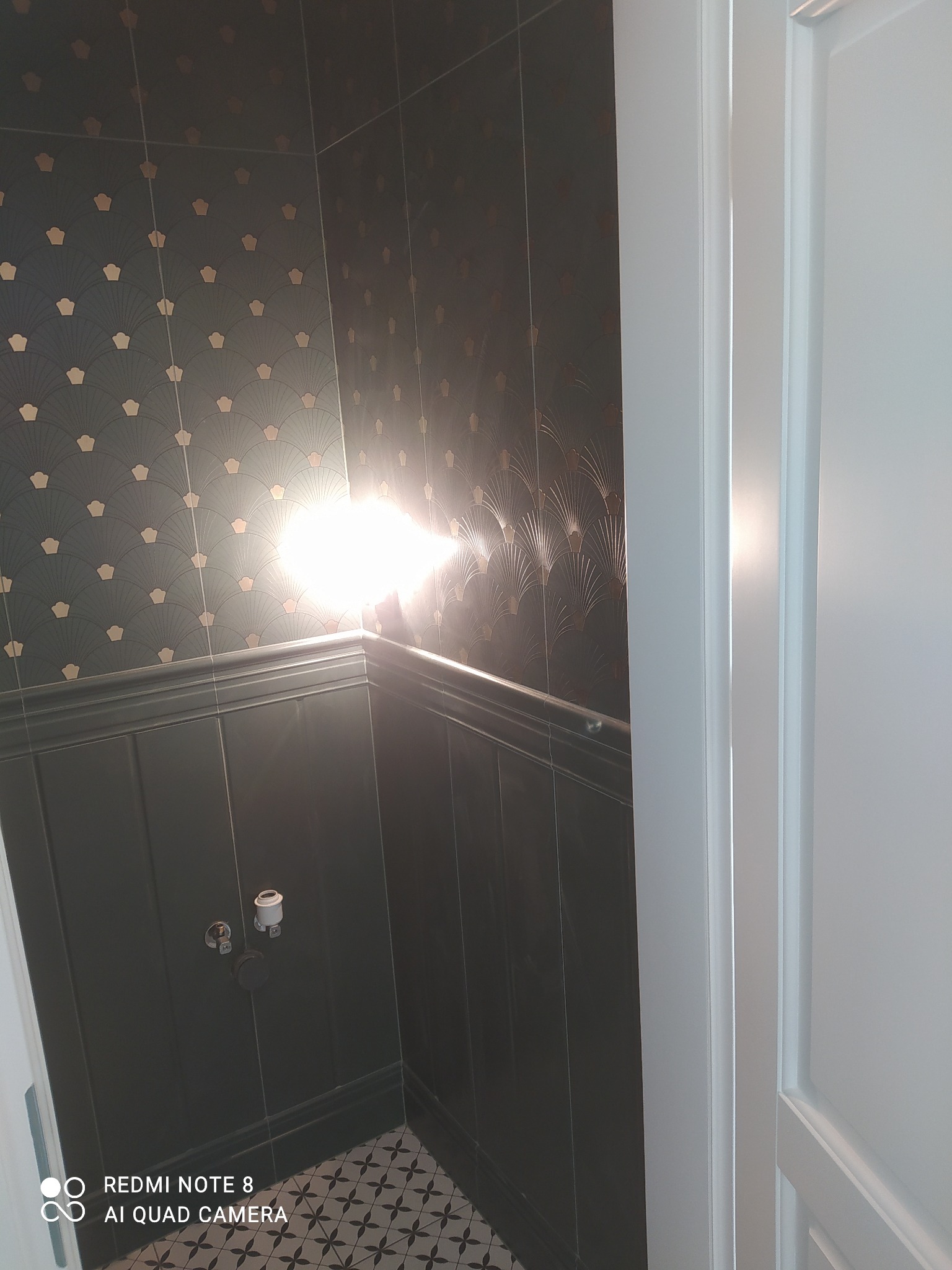 Ciemne płytki ścienne z geometrycznym wzorem w łazience, widoczne przyłącza wodne i fragment podłogi z wzorzystymi płytkami, biała ościeżnica drzwiowa po prawej stronie.