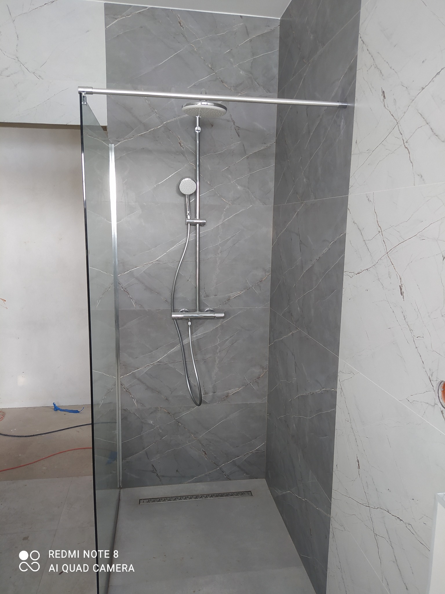 Szklana kabina prysznicowa typu walk-in z deszczownicą i odpływem liniowym, ściany wyłożone płytkami imitującymi marmur w odcieniach szarości i bieli, widoczne ślady prac wykończeniowych na podłodze.