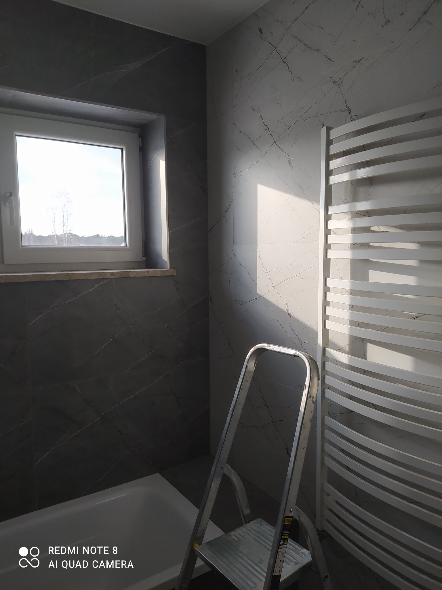 Łazienka w trakcie remontu z marmurowymi płytkami, widoczna drabina aluminiowa, grzejnik drabinkowy i wanna, oświetlona promieniami słońca wpadającymi przez okno.