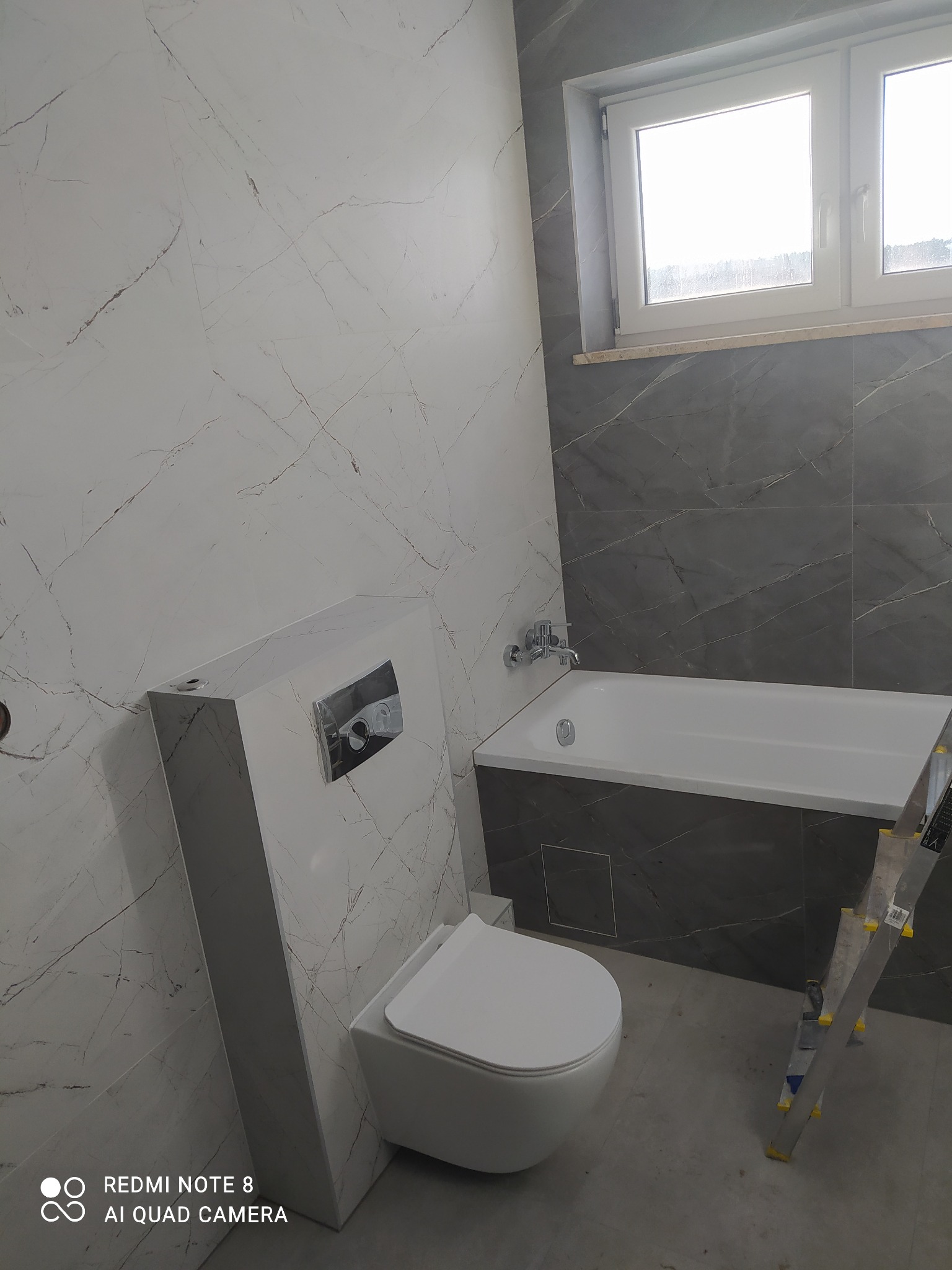 Nowoczesna łazienka w trakcie wykańczania: biała toaleta podwieszana z zabudową, wanna obłożona ciemnoszarymi płytkami imitującymi marmur, białe płytki ścienne z szarymi żyłami, drabina malarska...