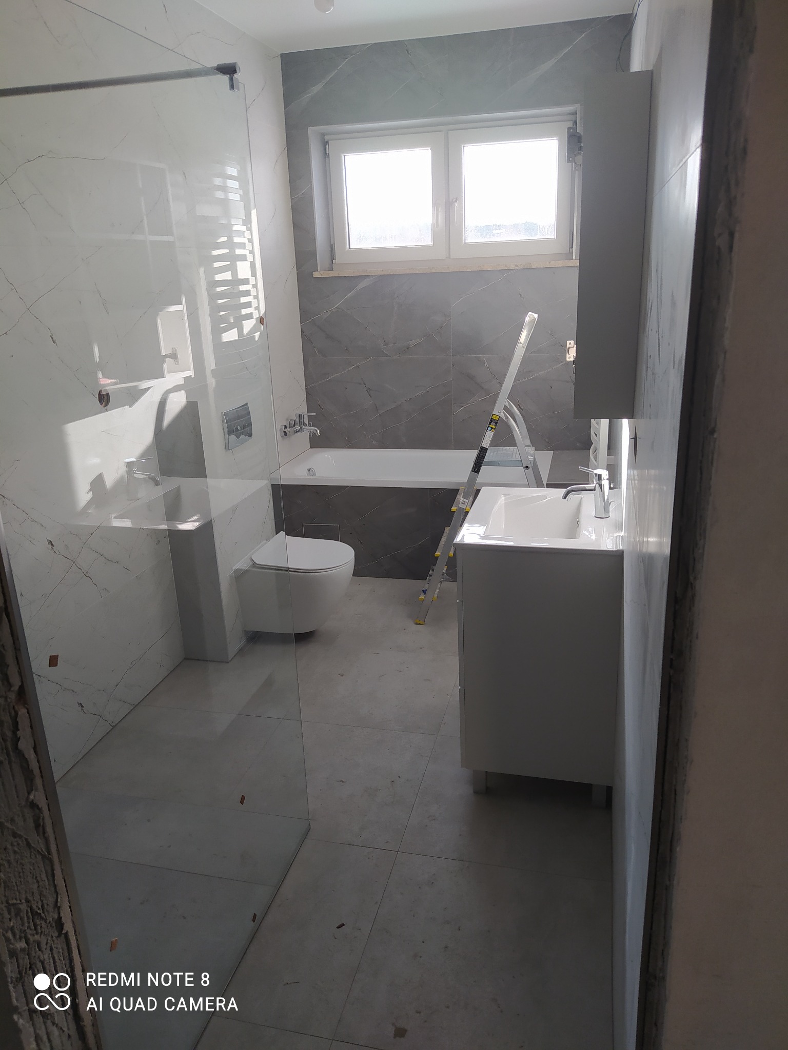 Wnętrze łazienki w trakcie wykańczania, widoczna kabina prysznicowa ze szklaną ścianą, umywalka z szafką, toaleta, wanna oraz drabina malarska.