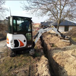 KOSPOL - BUD SPÓŁKA Z OGRANICZONĄ ODPOWIEDZIALNOŚCIĄ - Mini koparka Bobcat E19 w trakcie wykonywania wąskiego wykopu w piaszczystej glebie, widoczny operator w kabinie, w tle dom w trakcie budowy i drzewa bez liści.