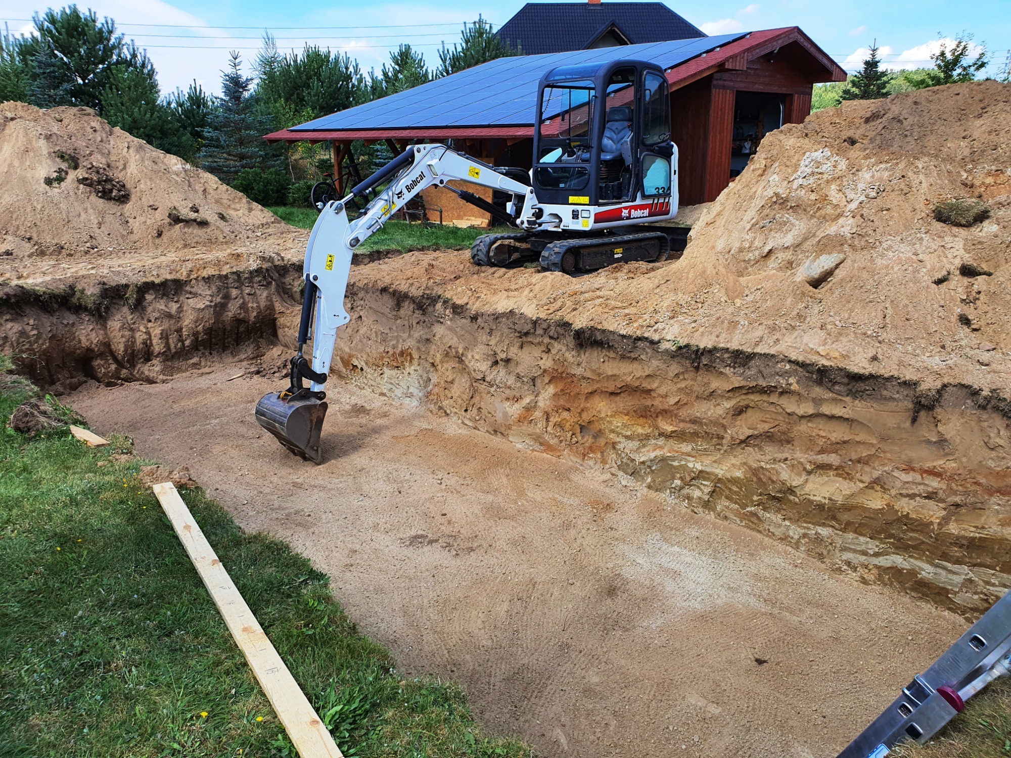 Mini koparka Bobcat w trakcie wykonywania wykopu pod fundamenty na trawiastym terenie, z widocznymi hałdami ziemi i drewnianą łatą pomiarową.