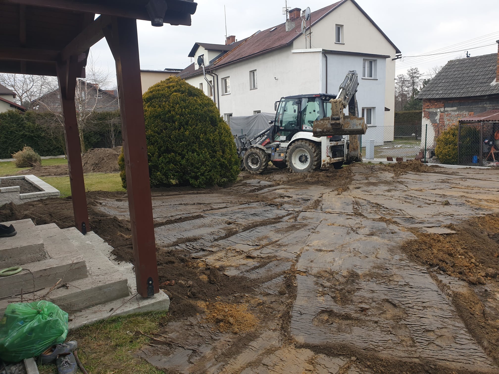 Biała koparko-ładowarka HOMEC w trakcie prac ziemnych na posesji, widoczne ślady opon na ubitej ziemi i pryzma ziemi obok tuji.