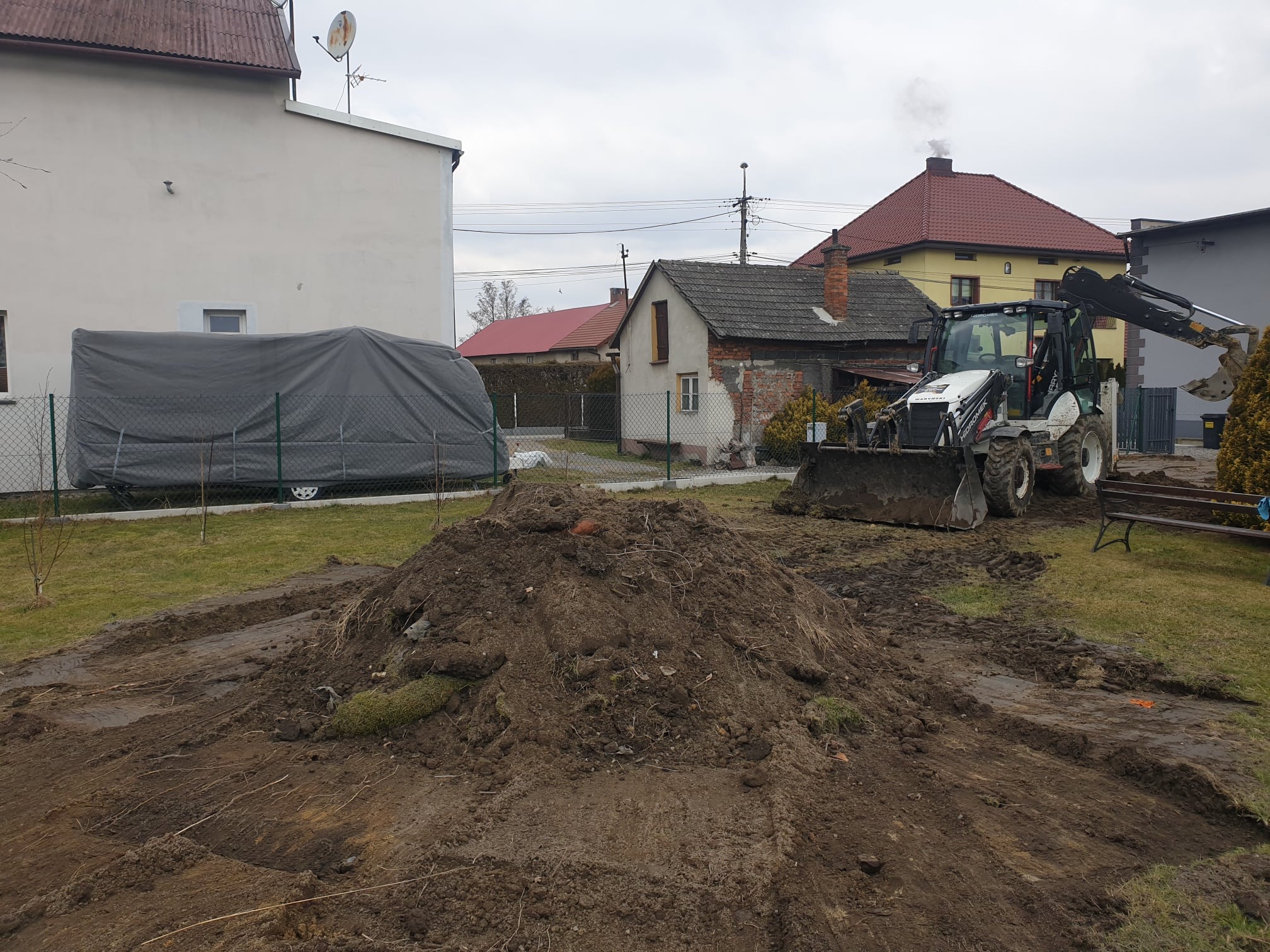 Koparko-ładowarka na działce, przygotowuje teren pod budowę, widoczny kopiec ziemi i ślady opon na błotnistym gruncie, w tle zabudowa jednorodzinna.