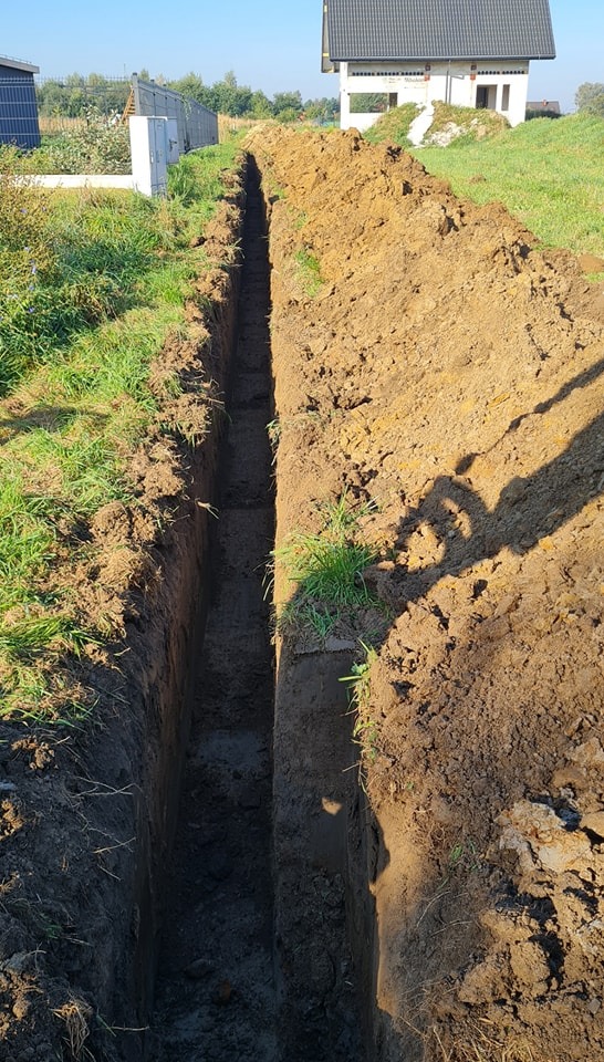 Długi, wąski wykop w ziemi, widoczny cień osoby robiącej zdjęcie, w tle budynek mieszkalny.