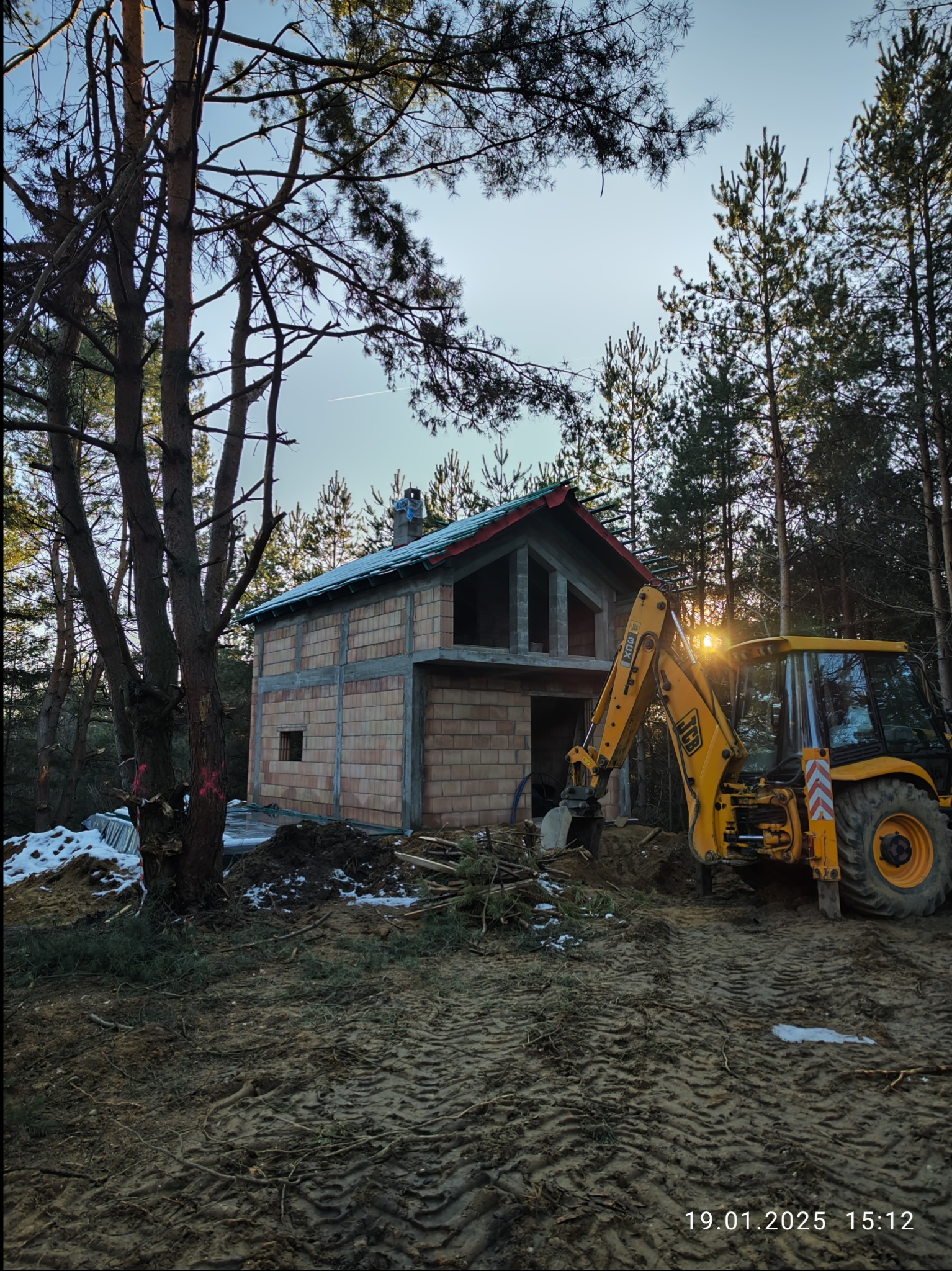 Niedokończony dom z pustaków otoczony lasem, z żółtą koparką JCB na pierwszym planie, przygotowującą teren. Na ziemi widać ślady opon i resztki śniegu.