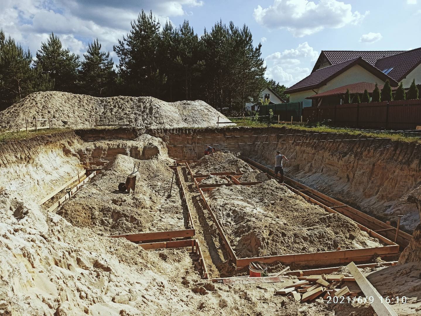 Wykop pod fundamenty domu jednorodzinnego z drewnianym szalunkiem, widoczny stos piasku i dwóch pracowników przy pracy.