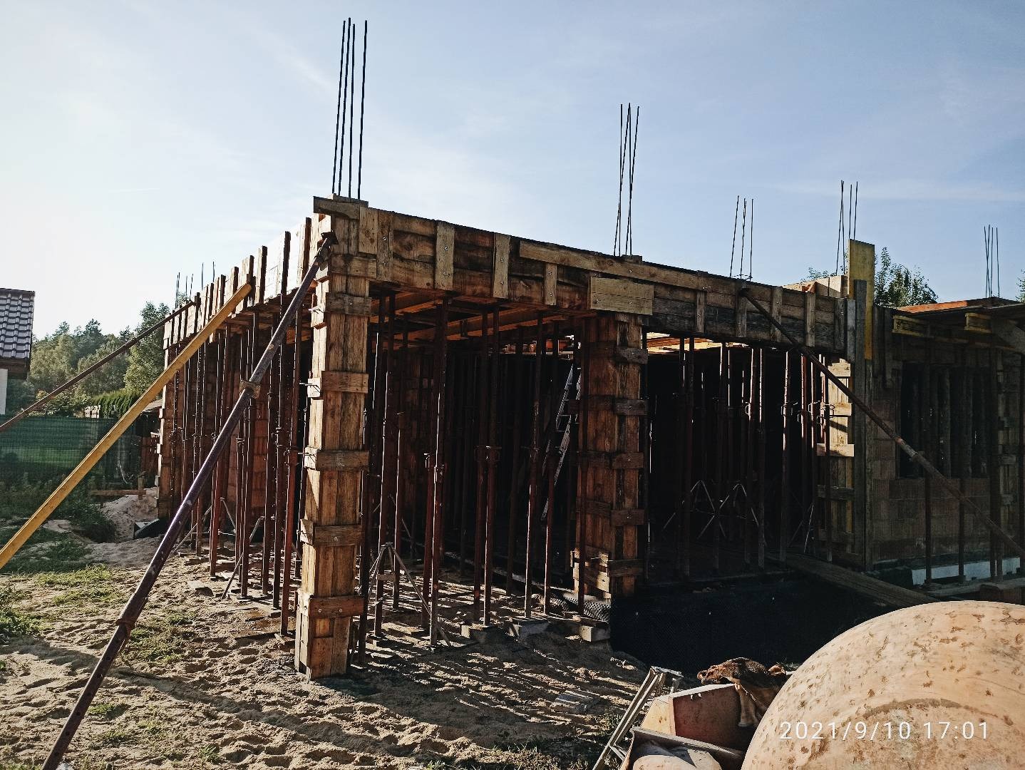 Surowa konstrukcja domu w trakcie budowy z drewnianym szalunkiem i wystającymi prętami zbrojeniowymi, widoczny piasek i częściowo zabetonowane fundamenty.