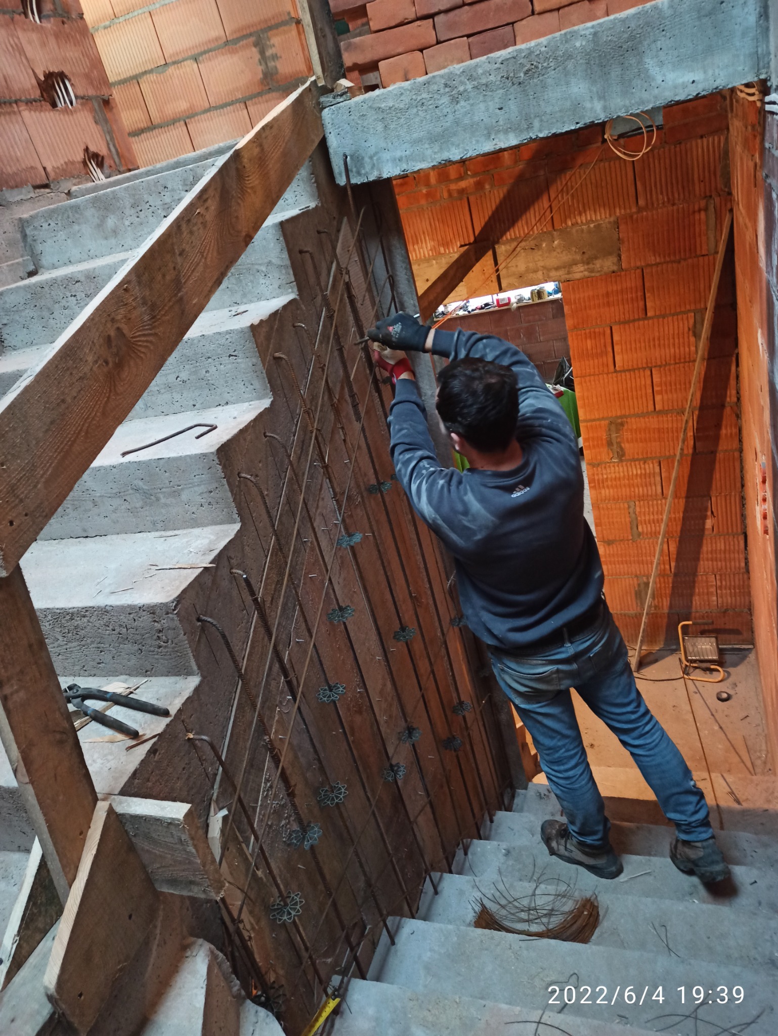 Pracownik w trakcie montażu zbrojenia do betonowych schodów wewnątrz budynku, widoczne ceglane ściany i tymczasowe oświetlenie.