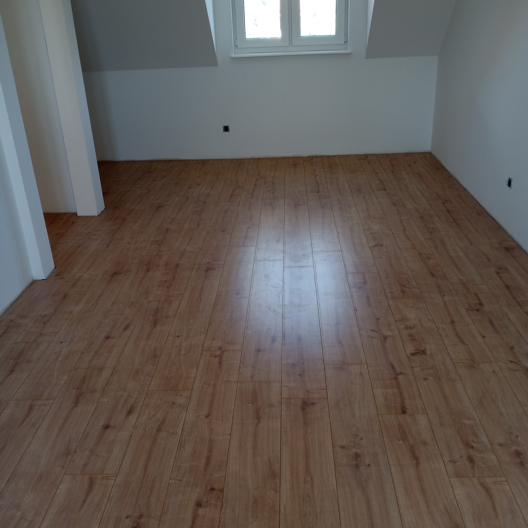 Ułożone panele podłogowe w jasnobrązowym odcieniu imitującym drewno, widoczne gładkie białe ściany i okno dachowe w wykończonym pokoju na poddaszu.