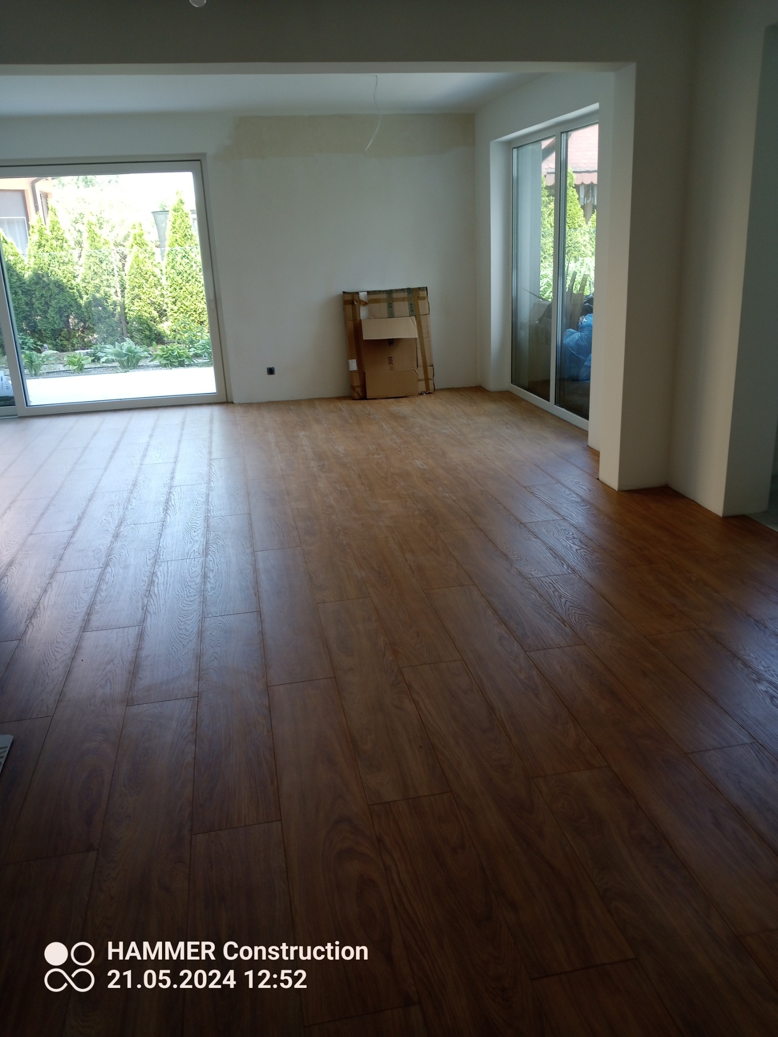 Ukończona podłoga z paneli w pomieszczeniu z dużymi oknami, widoczne kartony po panelach przy ścianie, fragment ściany przygotowany pod malowanie.