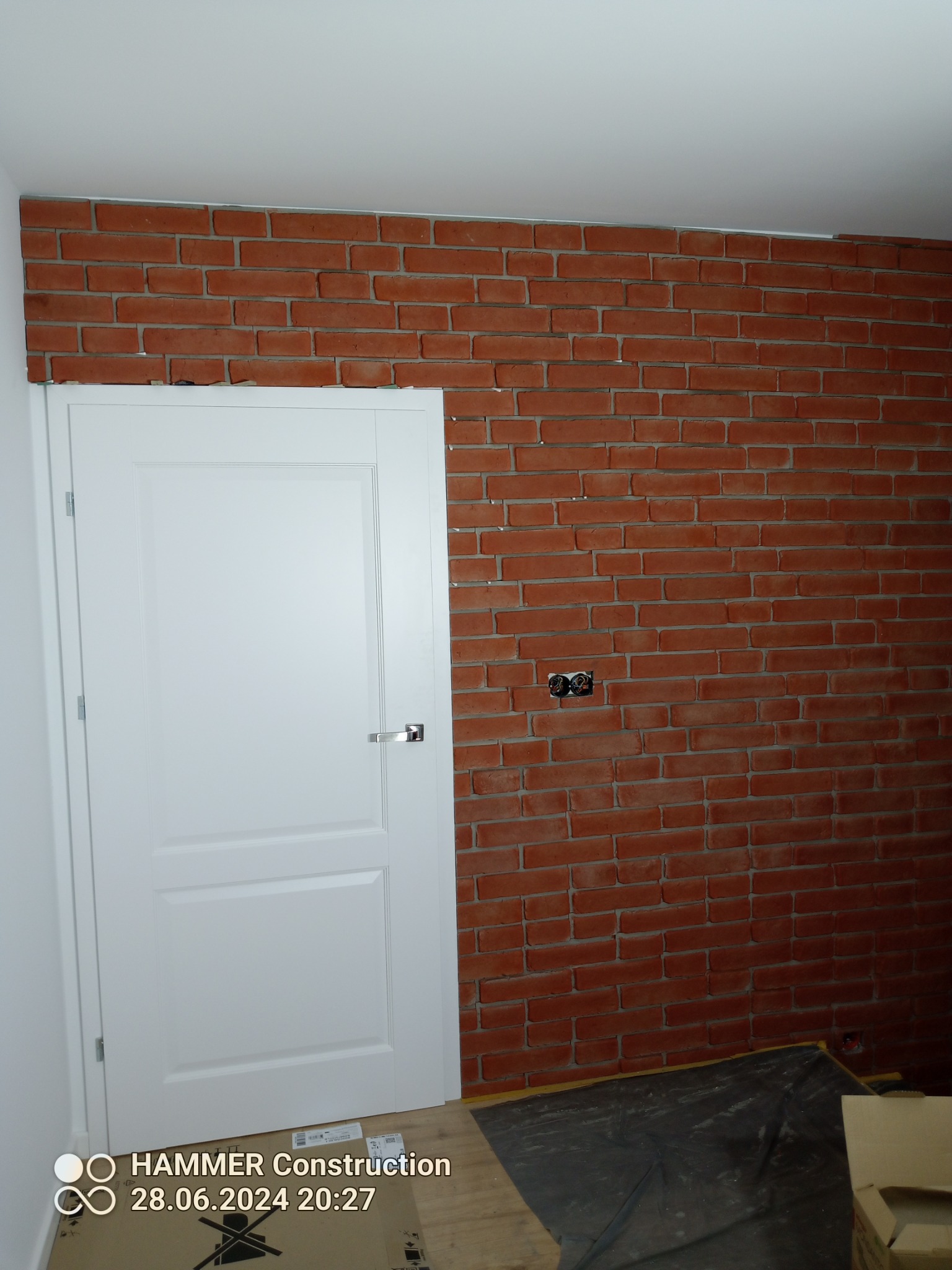 Ściana wykończona czerwoną cegłą klinkierową, z widocznym gniazdkiem elektrycznym i białą, klasyczną stolarką drzwiową.