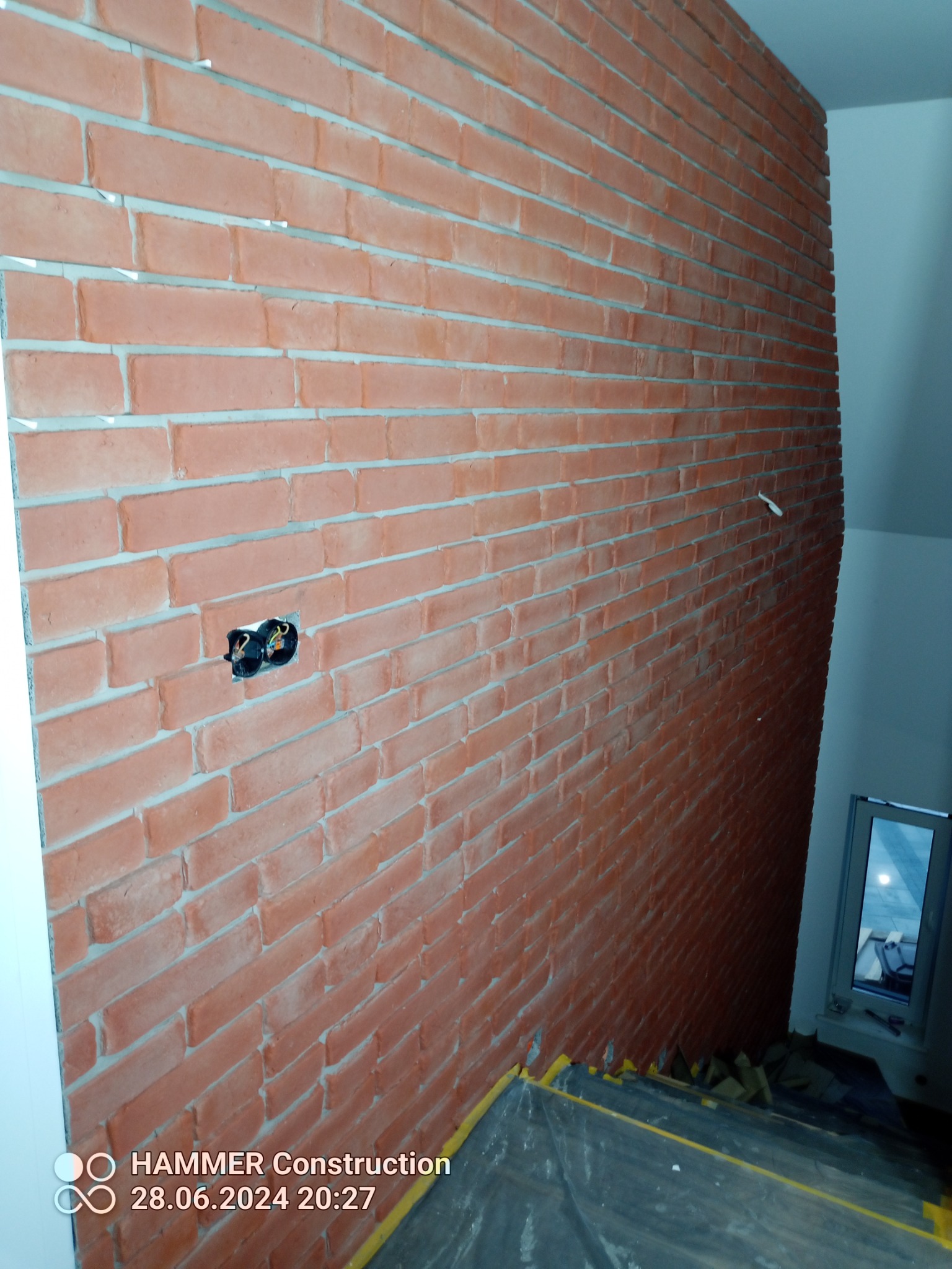 Ściana z cegieł klinkierowych z widoczną puszką elektryczną, wykończona jasną fugą, zabezpieczona folią malarską u dołu