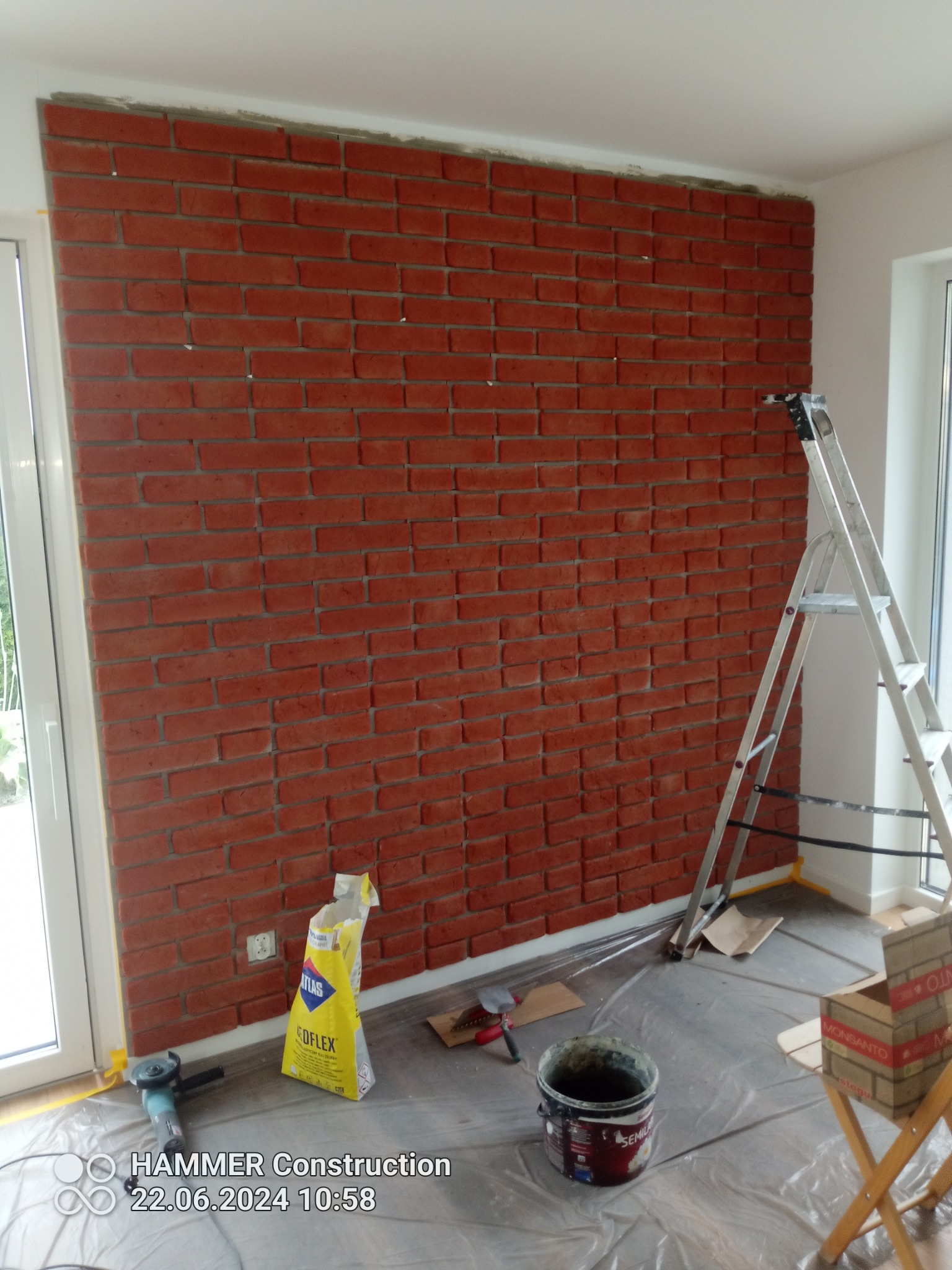 Ściana wykończona czerwoną cegłą dekoracyjną, widoczne narzędzia i materiały budowlane, folia ochronna na podłodze, drabina oparta o ścianę, etap prac wykończeniowych.