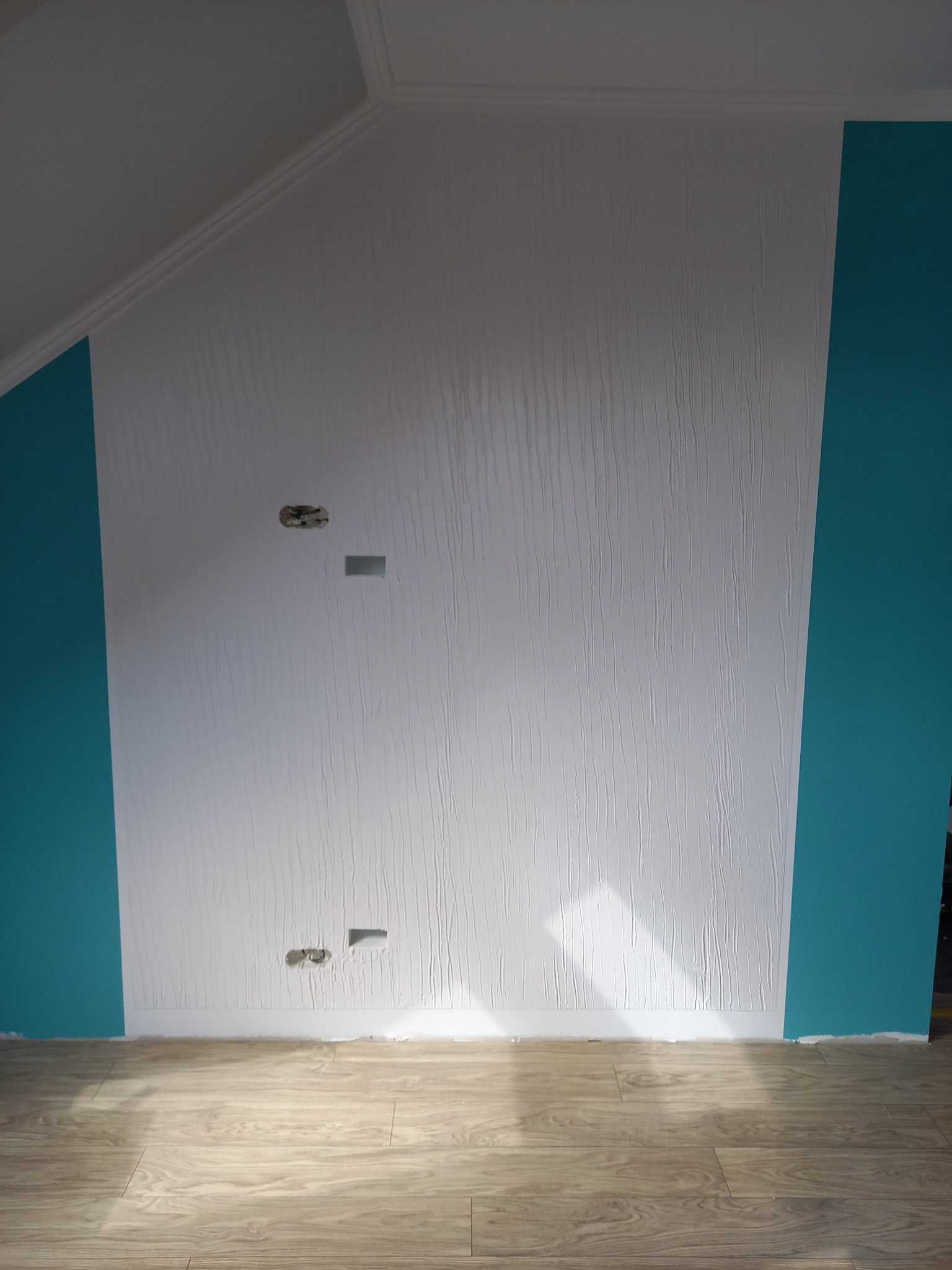 Biała ściana z fakturą, fragment turkusowej ściany, odsłonięte gniazdka elektryczne i panele podłogowe w pokoju na poddaszu.