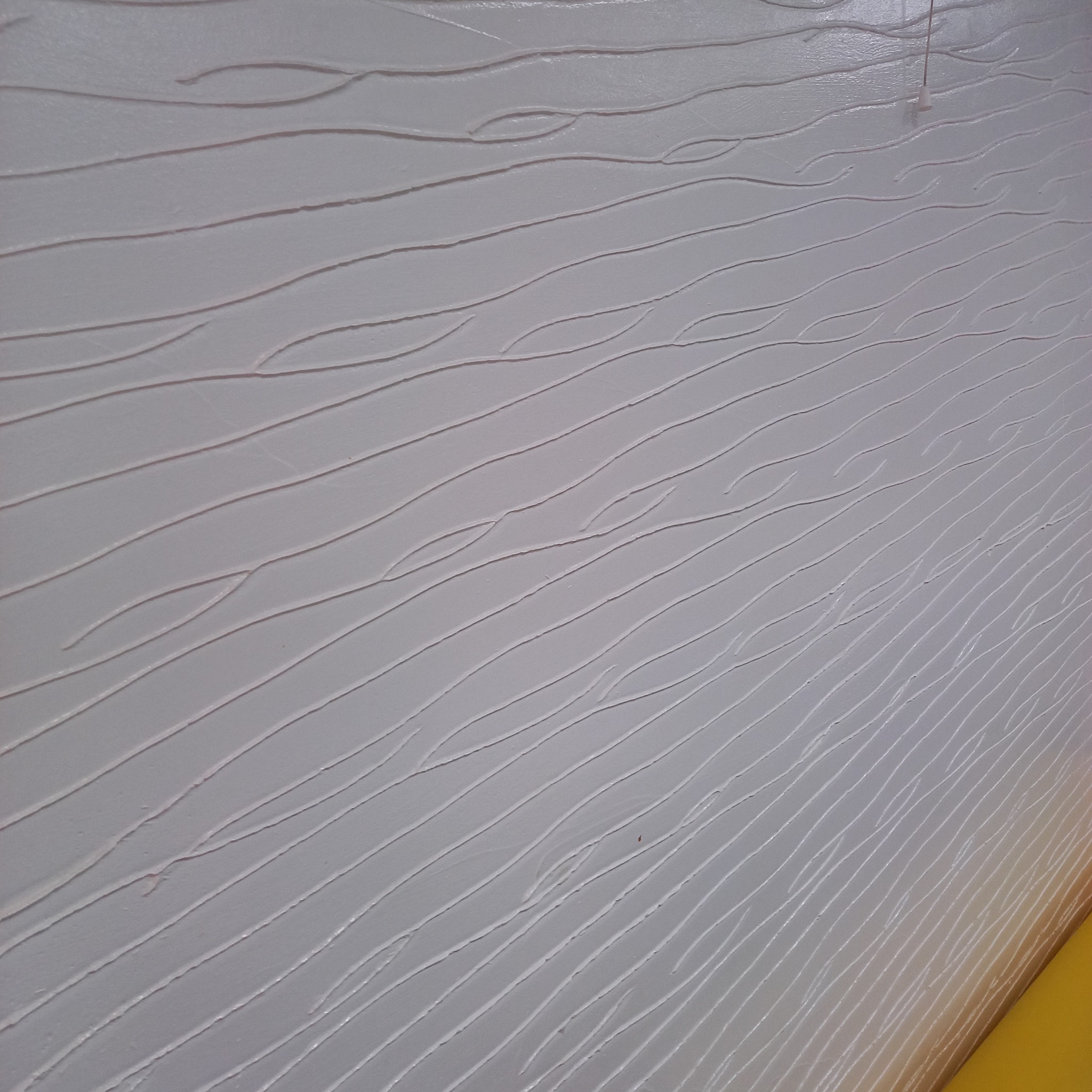 Biała ściana z dekoracyjną fakturą przypominającą poziome, nieregularne linie, oświetlona z góry, widoczny fragment żółtej listwy przypodłogowej.