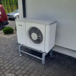 Biała pompa ciepła Stiebel Eltron zamontowana na metalowej konstrukcji na kostce brukowej, widoczny fragment elewacji budynku i zaparkowany samochód