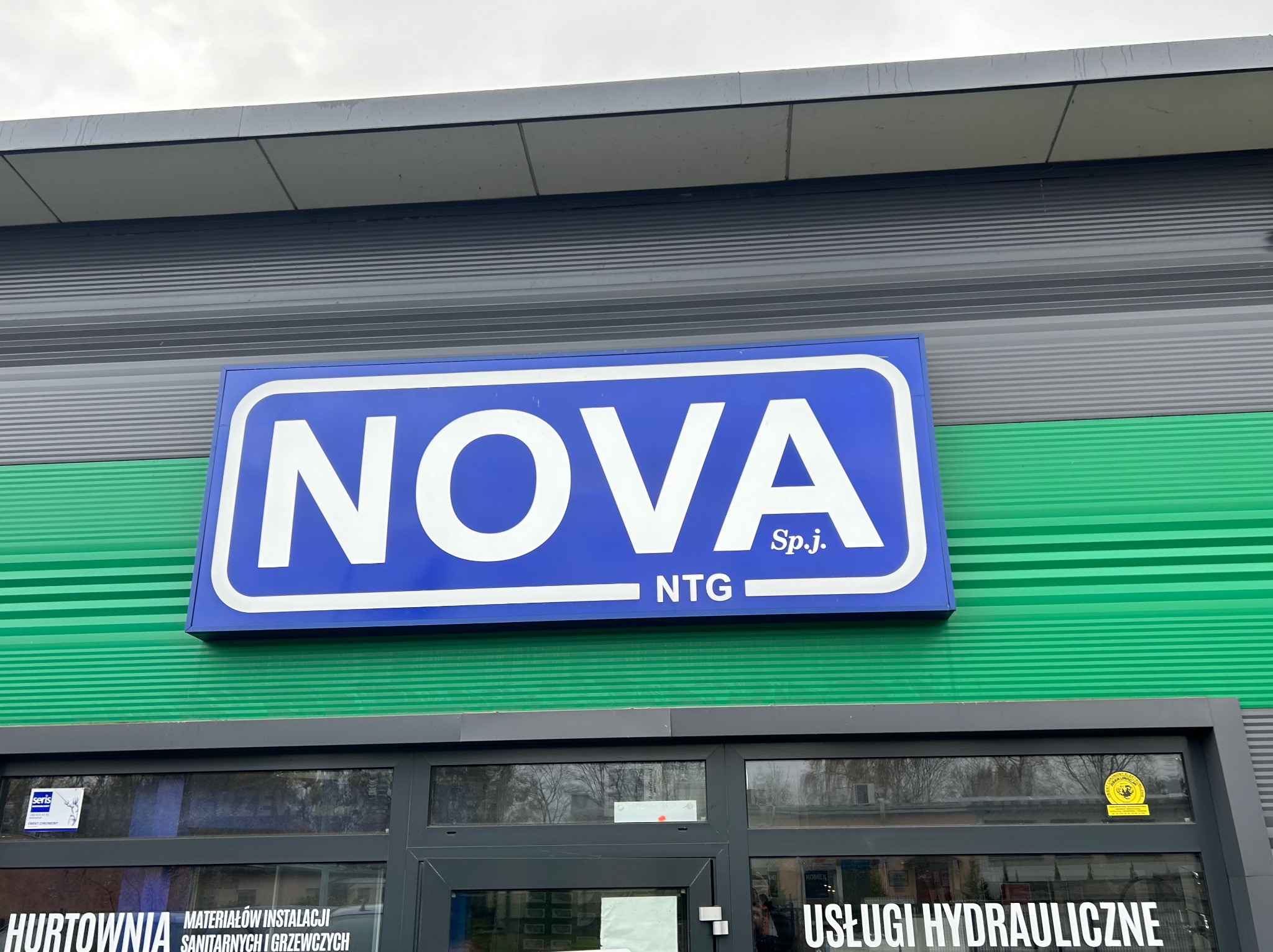 Zewnętrzny szyld firmy NOVA NTG Sp.j. oferującej usługi hydrauliczne i hurtownię materiałów instalacyjnych sanitarnych i grzewczych, widoczny nad wejściem do budynku z szarą fasadą i zielonymi...