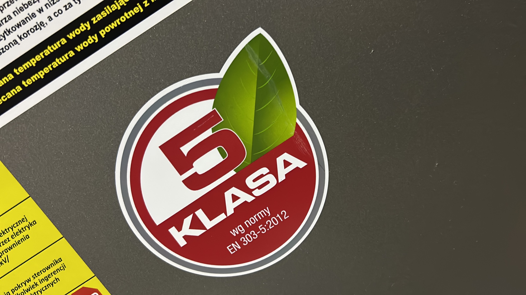 Etykieta '5 Klasa' z zielonym liściem, informująca o normie EN 303-5:2012, umieszczona na szarym urządzeniu grzewczym.