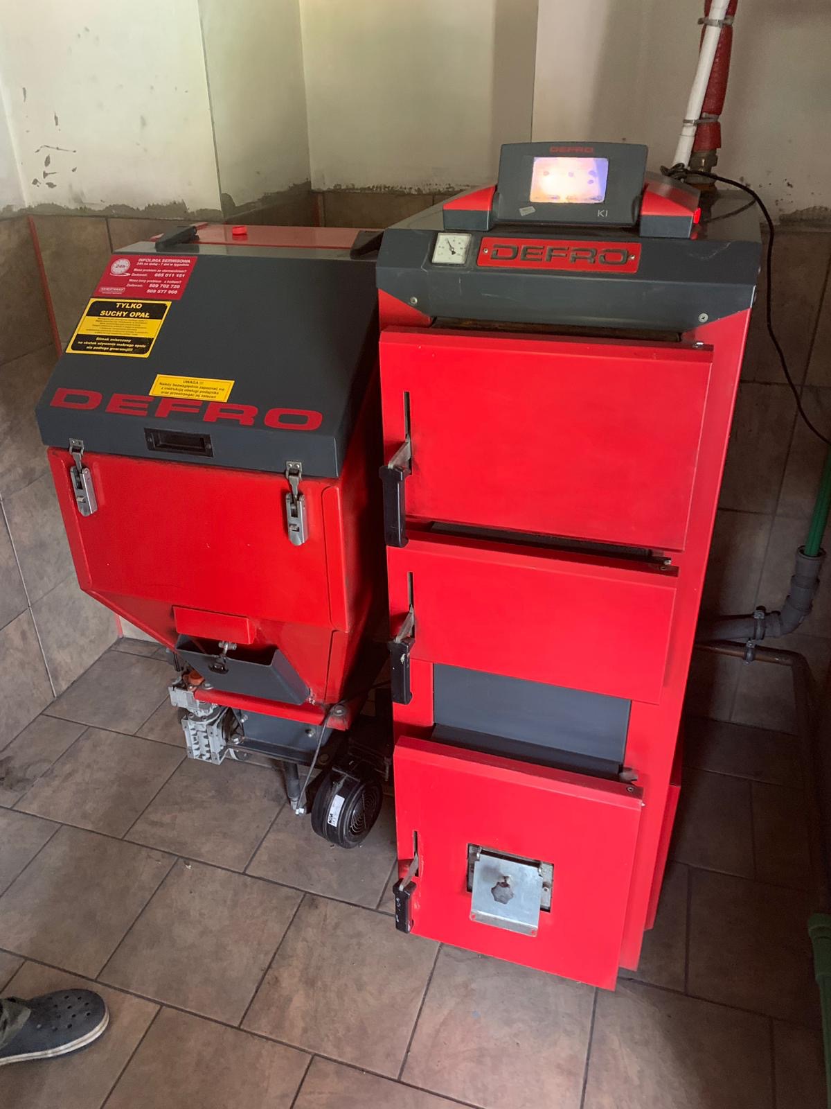 Czerwono-szary piec Defro z podajnikiem na ekogroszek, widoczny w piwnicy na brązowych płytkach, z fragmentem zielonej rury instalacyjnej w tle.