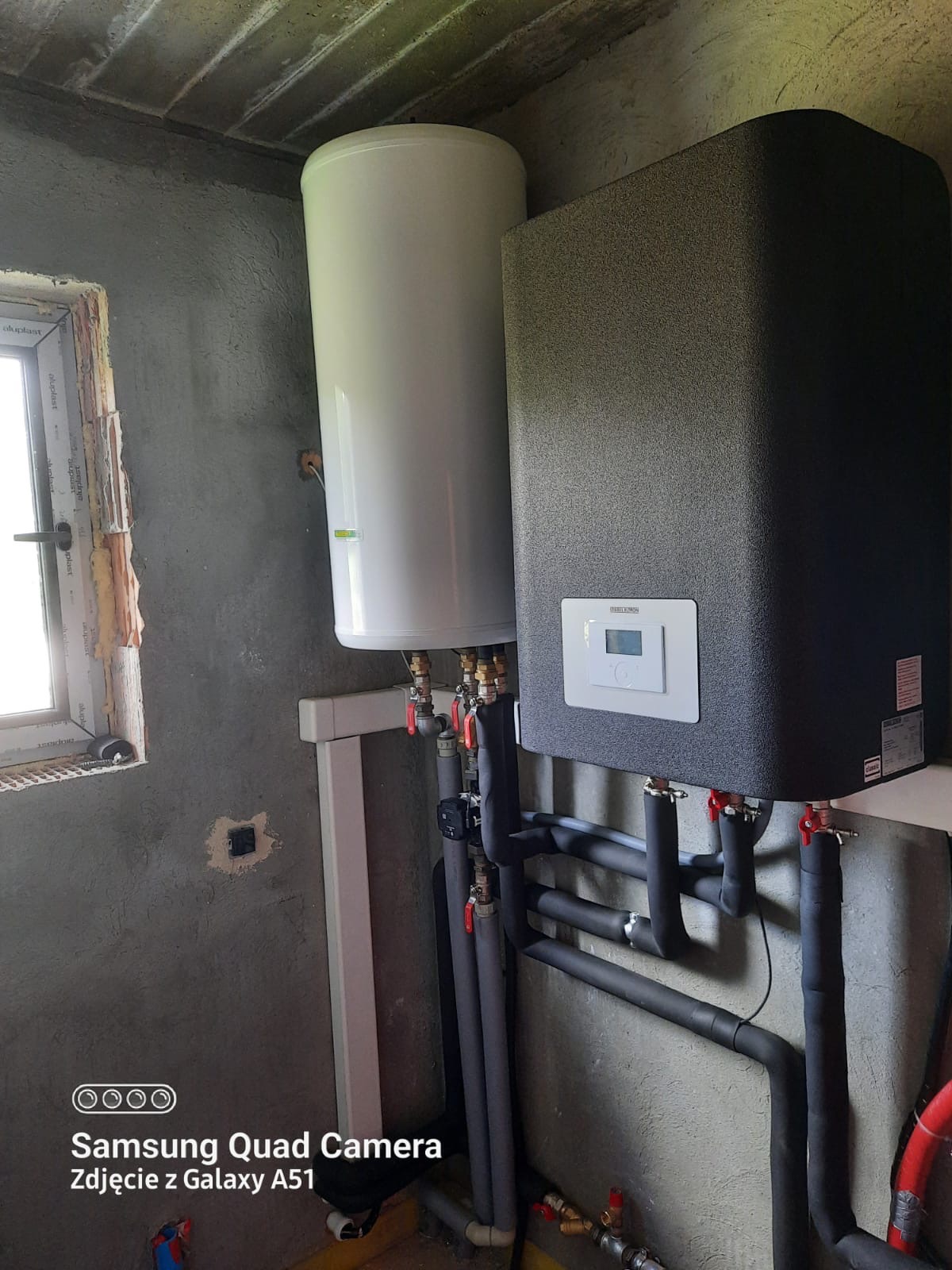 Kocioł grzewczy z zasobnikiem ciepłej wody, widoczne rury instalacji centralnego ogrzewania, surowy tynk na ścianie, okno z widocznymi śladami montażu.