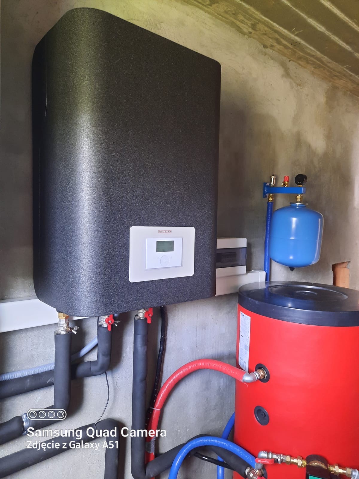 Czarny zbiornik buforowy ciepła marki Stiebel Eltron z podłączonymi rurami w izolacji, obok czerwony zasobnik CWU i niebieskie naczynie przeponowe w surowym pomieszczeniu.