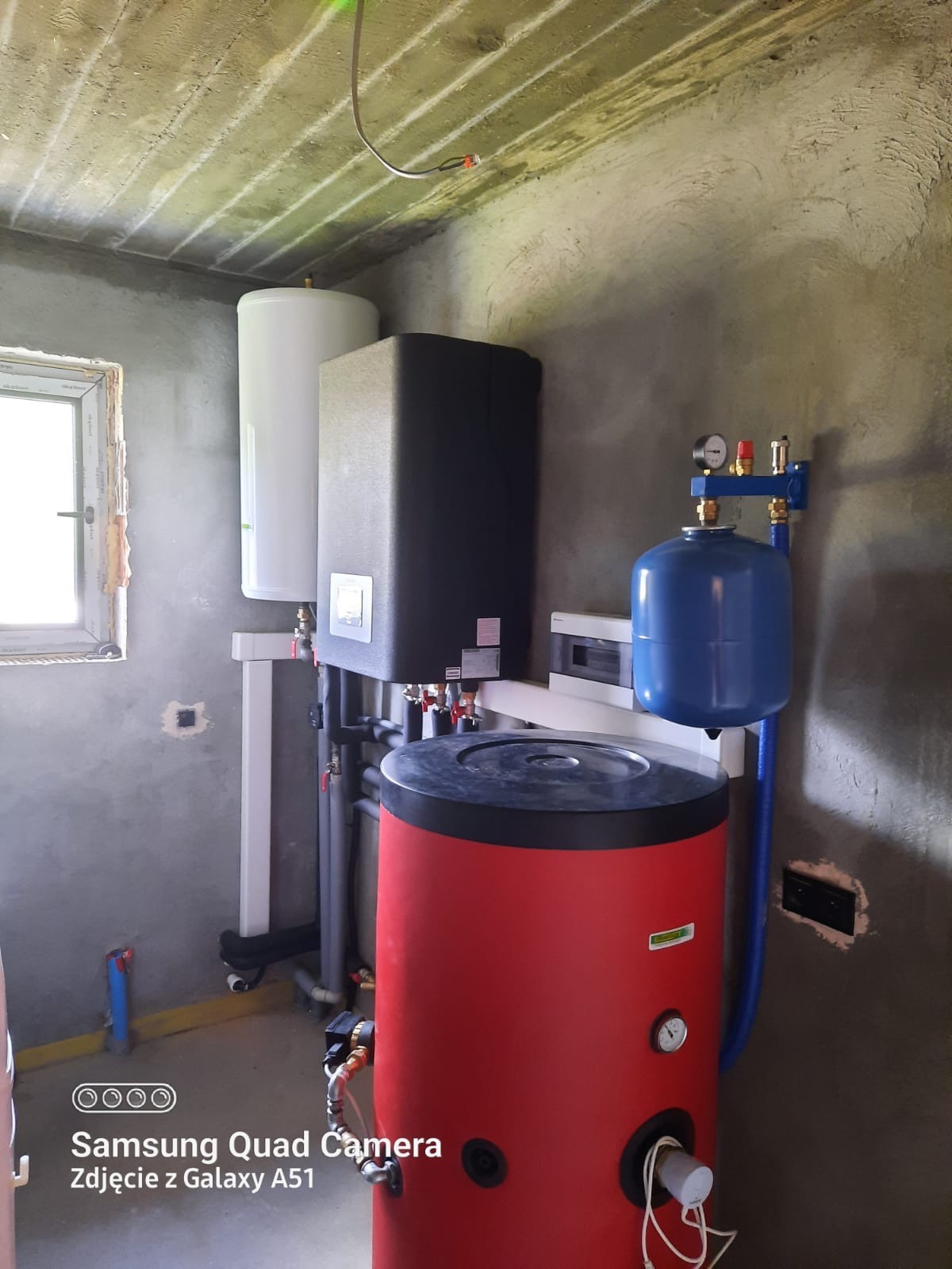 Kotłownia w surowym stanie: czerwony zbiornik buforowy, szary piec, biały zasobnik CWU, niebieskie naczynie przeponowe, rury instalacyjne, betonowe ściany i sufit.