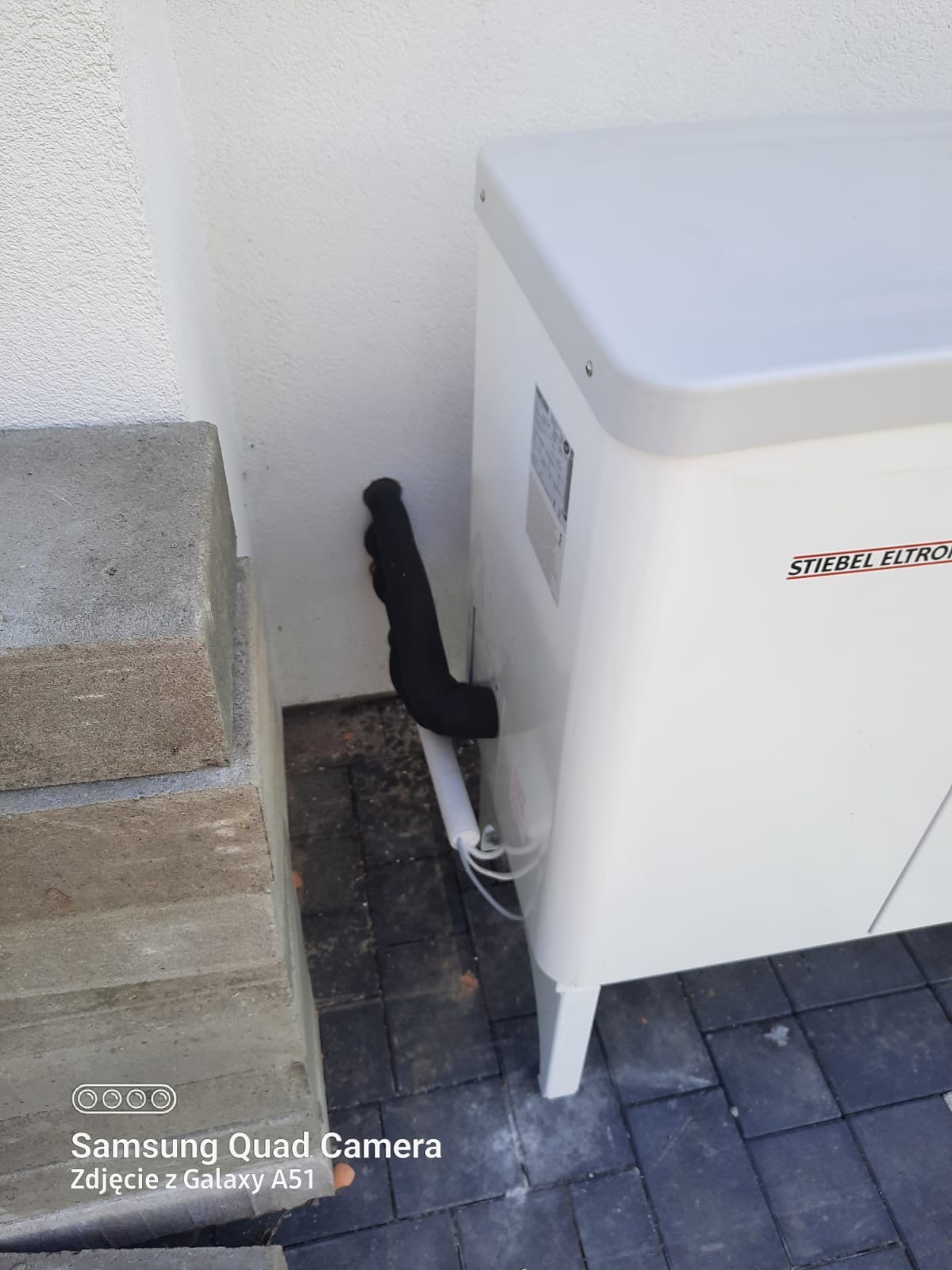 Biała pompa ciepła marki Stiebel Eltron zamontowana przy jasnej ścianie budynku, widoczne czarne rury izolacyjne i szara kostka brukowa.