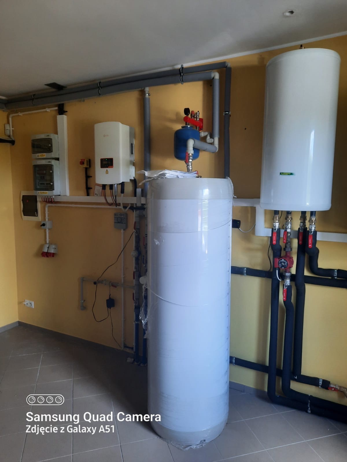 Bojlery i instalacja grzewcza w pomieszczeniu z żółtymi ścianami, widoczne rury, zawory, osprzęt elektryczny i zabezpieczony folią bojler.