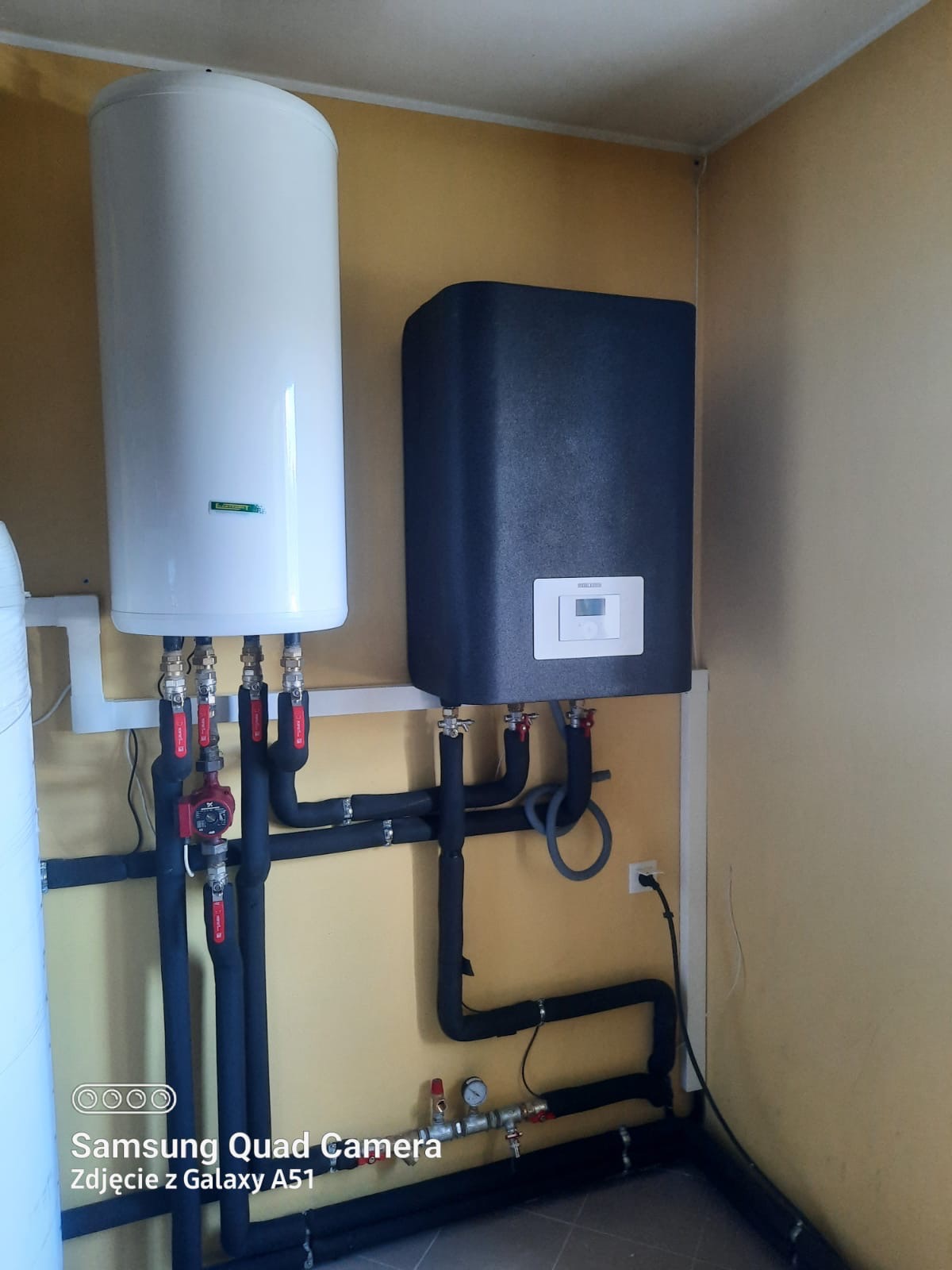 Biały bojler i czarny piec grzewczy zamontowane na żółtej ścianie, widoczne rury w izolacji, zawory i podłączenie do prądu.