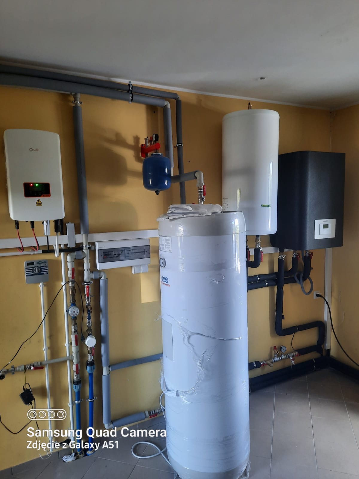 Kompletna instalacja grzewcza z zasobnikiem ciepłej wody owiniętym folią, inwerterem solarnym Solis, naczyniem przeponowym i licznikiem, zamontowana na żółtej ścianie.