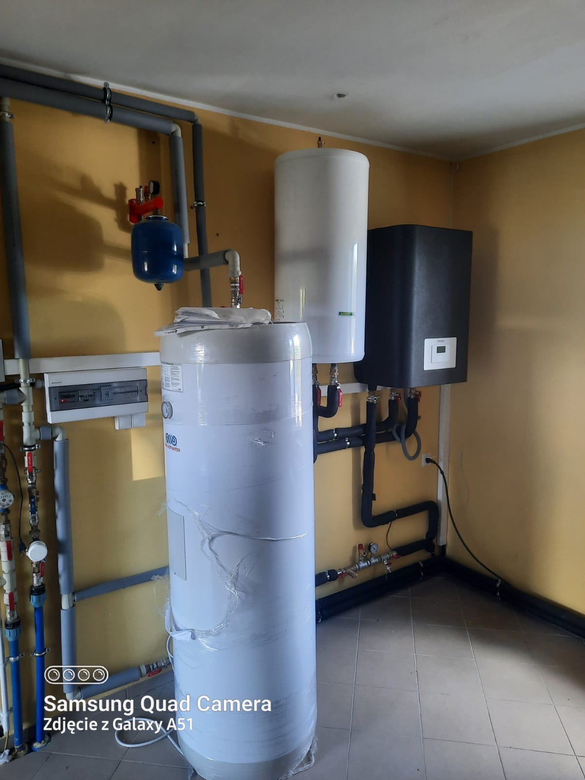 Nowa instalacja grzewcza w kotłowni: biały zasobnik ciepłej wody owinięty folią, czarny piec z wyświetlaczem, niebieskie naczynie wzbiorcze i szare rury na tle żółtych ścian.