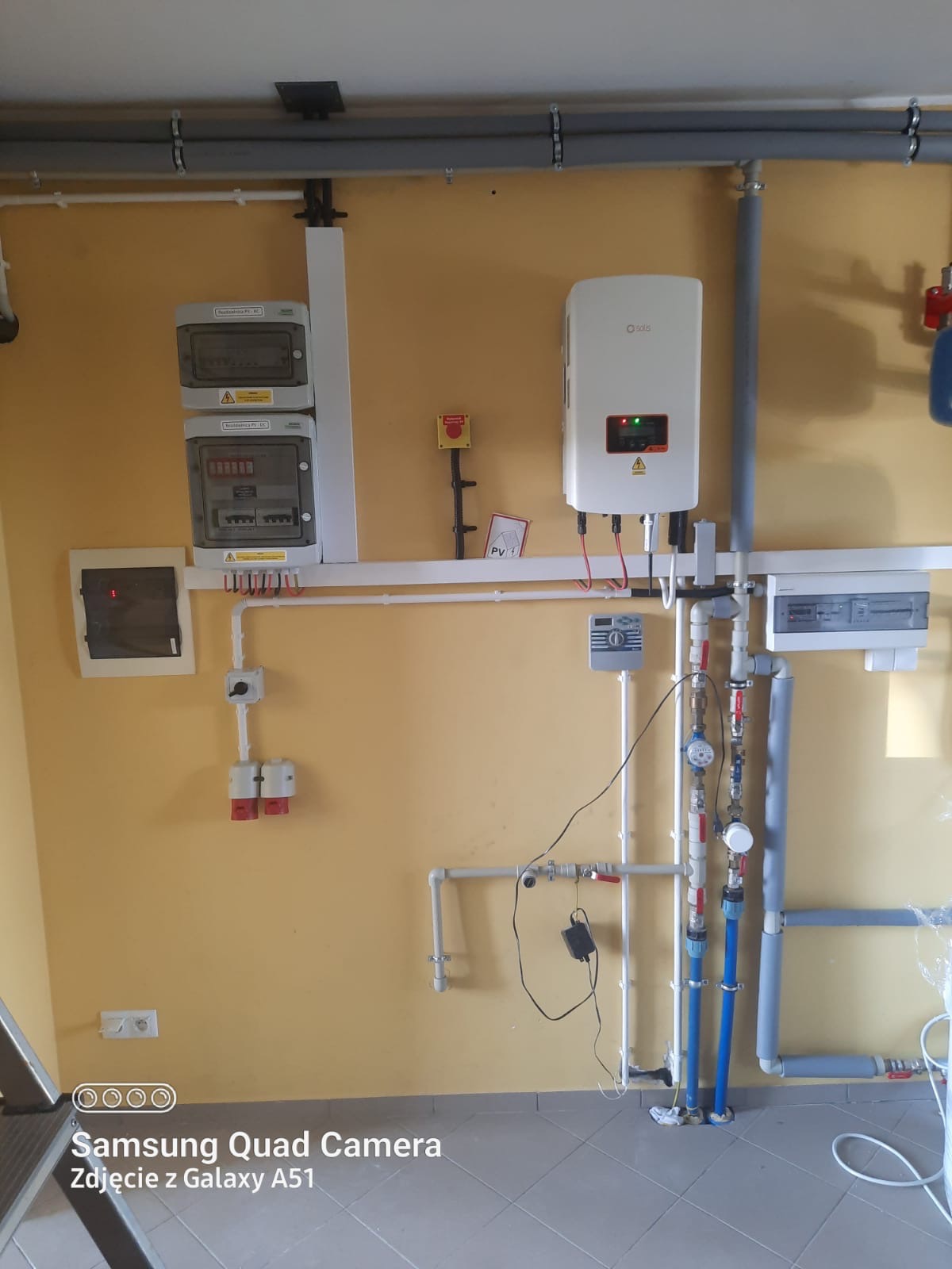 Biały inwerter solarny Solis zamontowany na żółtej ścianie, obok liczników elektrycznych i instalacji hydraulicznej z niebieskimi rurami, widoczne przewody elektryczne i rury PCV.